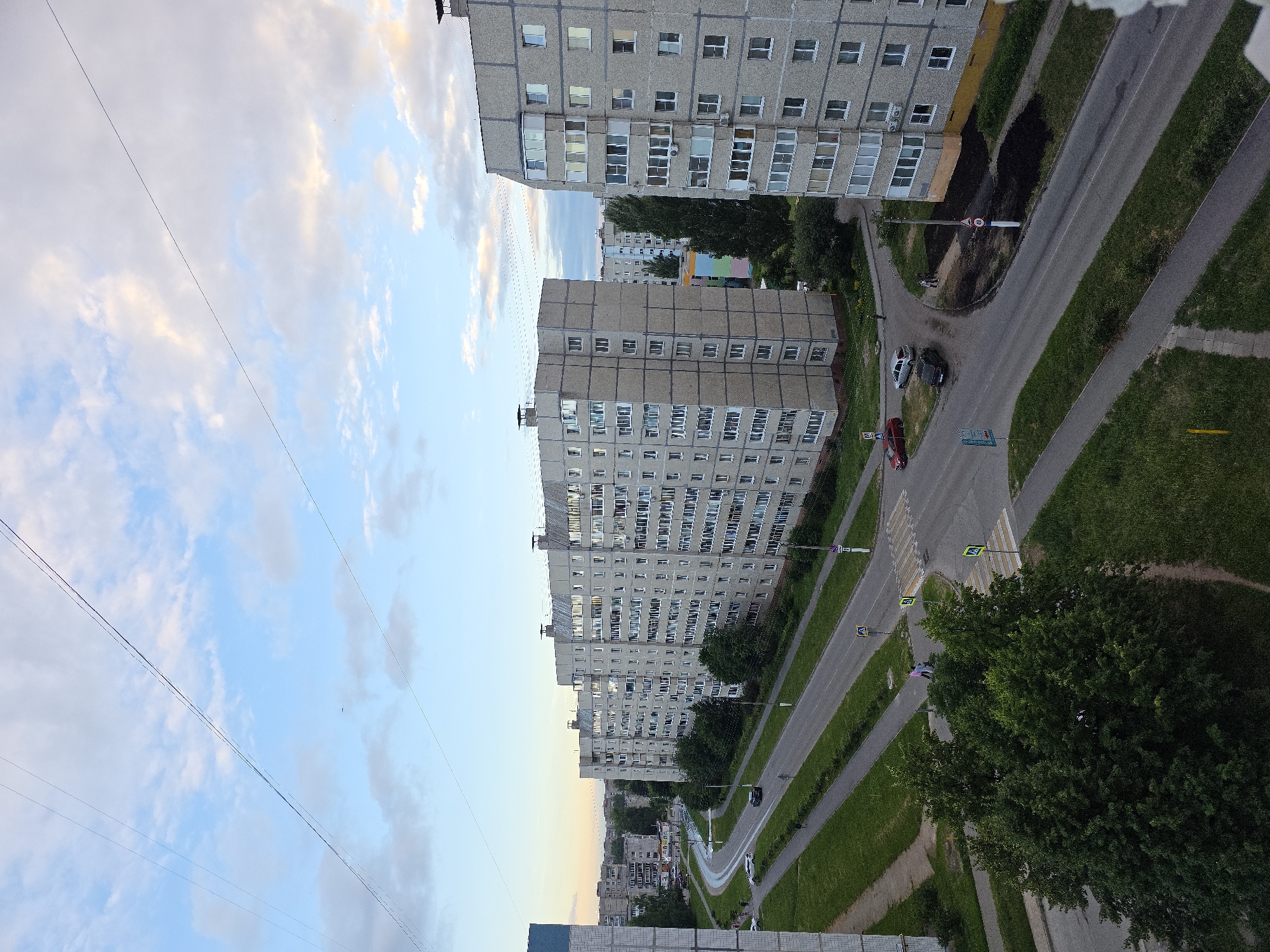An aerial view captures a city scene in Novocheboksarsk, Russia, under a partly cloudy sky. The image features several multi-story, concrete apartment buildings with numerous windows. A road with white lane markings curves through the scene, bordered by green grass and trees. Cars are visible on the road and parked. Pedestrians are present on sidewalks and at a crosswalk. Power lines stretch across the sky, and streetlights and traffic signs are also visible. The perspective suggests the photograph was taken from a higher vantage point, possibly another building.