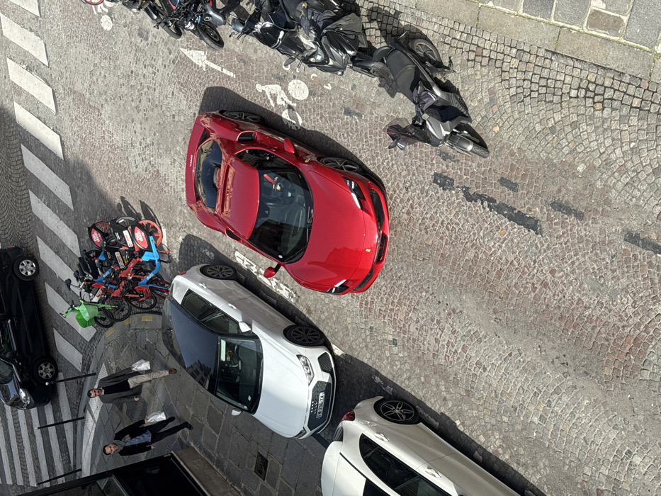 This image presents an elevated, angled view of a cobblestone street in Paris, France, under daylight conditions.
A prominent red Ferrari sports car is parked on the right side of the street, angled slightly towards the upper left. Behind it, a white Audi A1 hatchback with a black roof is parked, displaying the French license plate "FR 236 YW". A partial view of a third white compact car is visible on the far left. Near the top of the frame, a black car is present adjacent to a white pedestrian crossing. A cluster of shared bicycles, including green, blue, and red models, are parked on the left side of the street. On the right, a row of multiple motorcycles and scooters is parked. A white bicycle symbol is painted on the cobblestone surface near the parked motorcycles.
Two adult males are walking along the left side of the street, near a dark building facade. The lead man wears a dark jacket and carries a white plastic bag in his left hand. The man following him wears a light-colored coat and carries two white plastic bags, one in each hand. Sunlight casts distinct shadows across the cobblestone pavement.