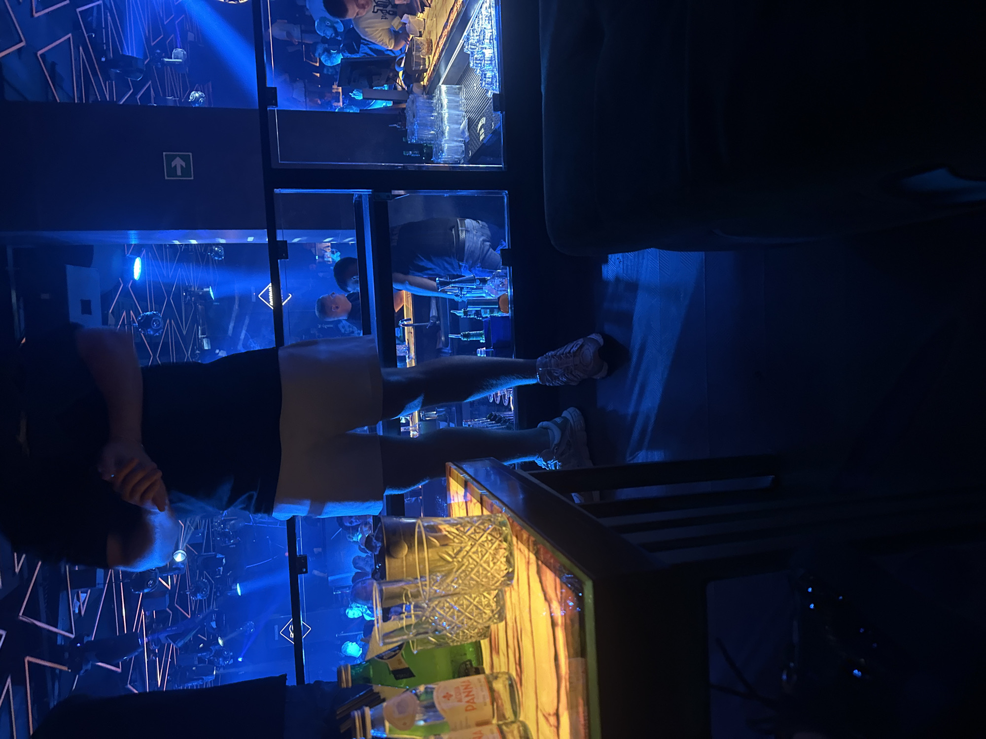 The image depicts the interior of a nightclub in Ruse, Bulgaria. The environment is dimly lit with predominantly blue and neon lighting, characteristic of a club setting. In the foreground, a person wearing a dark t-shirt and light-colored shorts stands with their hands clasped. To their left, a table displays several glass bottles of water, including one labeled "ACQUA PANNA," and a few empty patterned glasses.
In the background, through a glass partition, a bar area is visible. Bartenders and patrons are present. One bartender, wearing a dark shirt, is seen behind the bar, interacting with customers. Further back, other patrons are gathered, some standing and some seated at the bar. The club's ceiling features intricate geometric light fixtures, contributing to the overall ambiance. A green directional sign pointing to the right is affixed to a dark blue pillar. The overall impression is that of a lively nightlife venue.