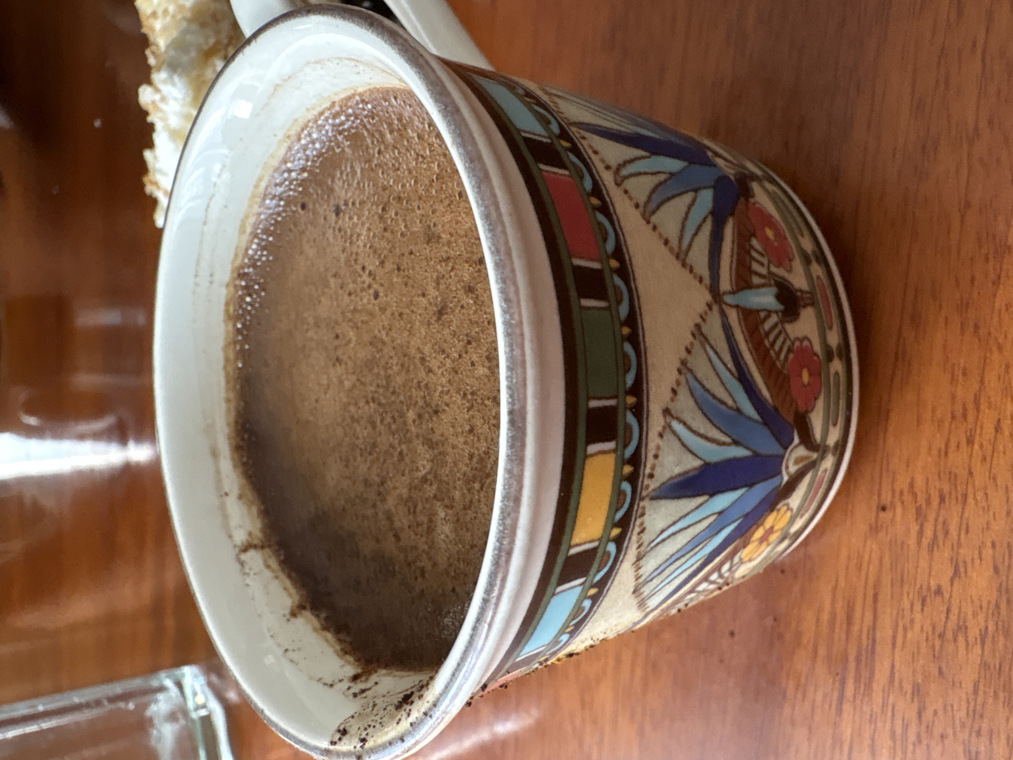 A close-up, eye-level shot captures a decorative mug filled with dark, frothy coffee on a wooden table. The mug, cream-colored with a band of colorful geometric patterns and floral motifs in blues, greens, reds, and yellows, sits slightly off-center. The coffee's surface is a rich brown with a fine foam. To the left of the mug, the blurry outline of a clear glass is visible, and to the right, a portion of a light-colored, pastry-like food item is out of focus. The table surface is a warm, polished wood with visible grain. The setting is a casual indoor environment. There is no indication of the specific location within Nha Trang Ward, Vietnam.