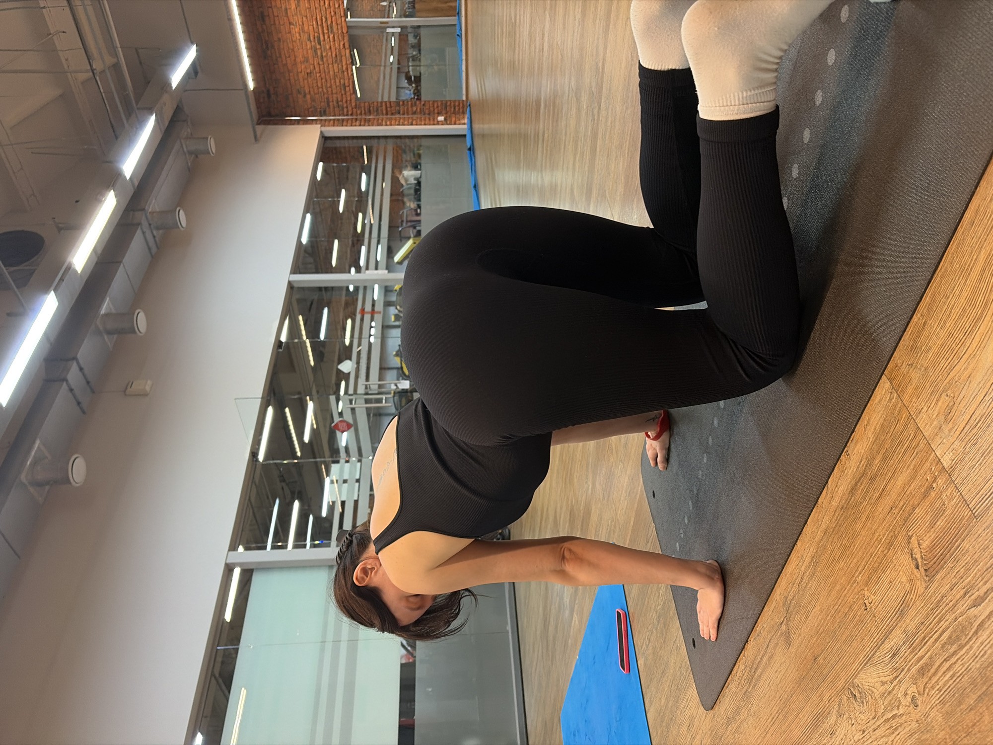 The image shows a woman practicing yoga on a mat in what appears to be a gym or studio. She is wearing black athletic wear, including leggings and a tank top, and white socks. The woman is in a kneeling position, with her hands on the mat and her upper body bent forward. The setting includes wooden flooring, a yoga mat, and a blue mat underneath. In the background, there are brick walls and large windows that reveal glimpses of gym equipment and other people. The lighting suggests it is daytime, possibly indoors with artificial lighting. The overall mood seems focused and calm, indicative of a yoga practice. There is no visible text in the image.