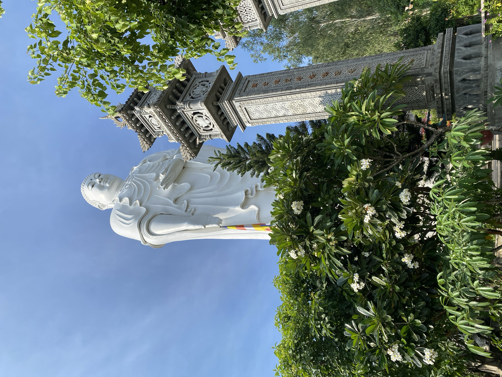 The image shows a tall, white statue of Buddha, seen from a low angle, against a clear blue sky. The statue is adorned with a colorful, striped flag hanging from its left side. In the foreground, lush green trees and flowering bushes partially obscure the lower part of the statue and two intricately carved stone pillars. The pillars are ornate, featuring geometric patterns and decorative elements. The overall scene suggests a peaceful and spiritual environment, likely a temple or religious site. The location is identified as Tay Nha Trang Ward, Vietnam.
