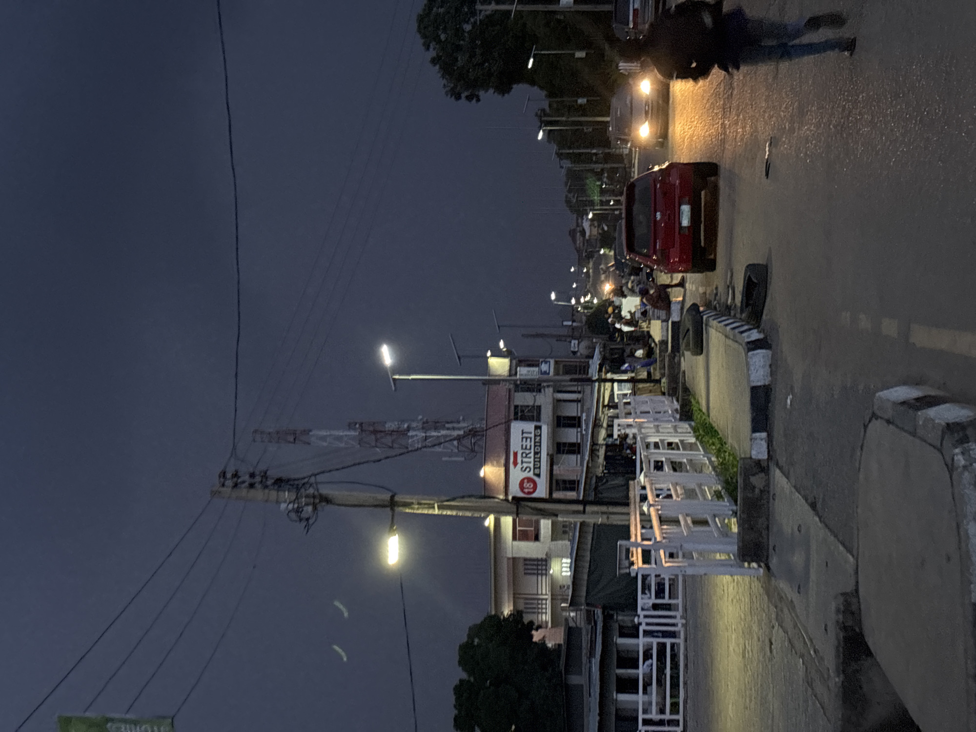 The image depicts a street scene at dusk in Jos, Nigeria, likely along a main thoroughfare. Overhead power lines crisscross the dark, overcast sky, with a prominent utility pole featuring multiple wires and insulators on the left side of the frame. A brightly lit street lamp illuminates the area around the pole. In the background, two tall communication towers, one red and white, are visible.
A building with a sign that reads "18° STREET BUILDING" with a red arrow pointing left is situated on the right side of the street. Several cars are on the road; a red hatchback is in the foreground, its brake lights illuminated. Another car with its headlights on is further down the street. Several people are visible on the sidewalks and near the road, some appearing to be pedestrians and others possibly street vendors.
The road surface appears wet and reflective, suggesting recent rain or damp conditions. A white railing runs along the side of the road, separating it from a paved area with some greenery. The overall atmosphere is dim and somewhat somber, characteristic of twilight in an urban environment. The presence of multiple streetlights suggests this is a well-trafficked area.
