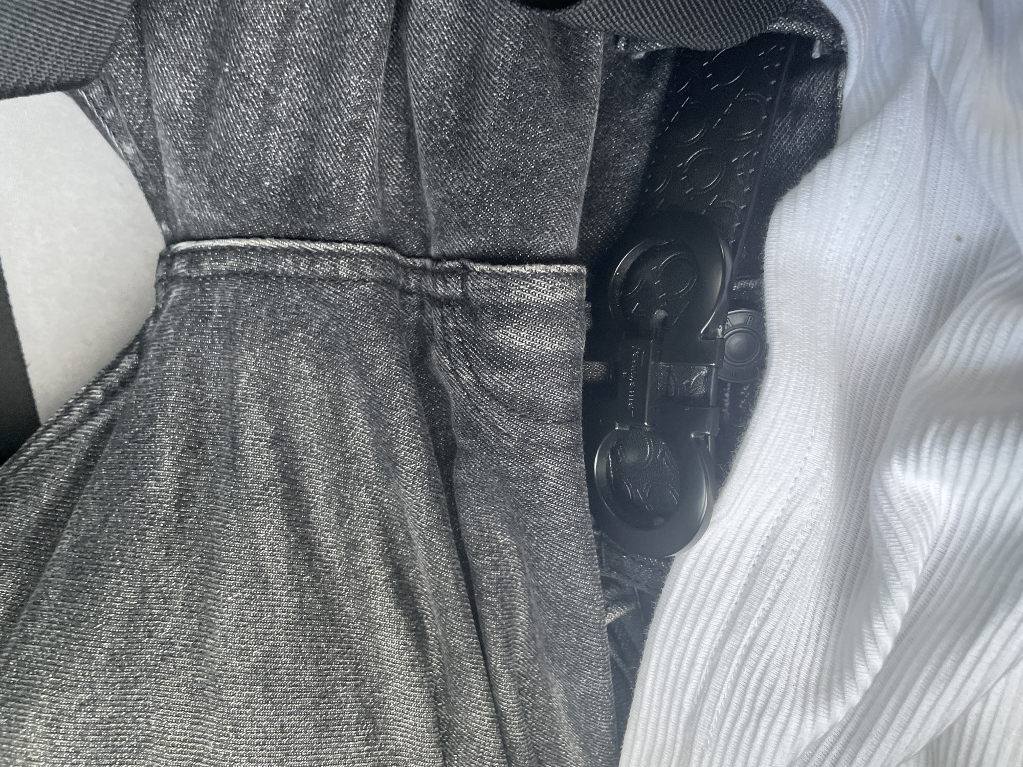 The image is a close-up, high-angle, eye-level shot of a black belt with a double G logo buckle, a pair of dark gray jeans, and a white ribbed t-shirt. The belt is fastened and threaded through the belt loops of the jeans. The jeans are made of denim with visible stitching. The t-shirt is folded beneath the belt and jeans, with its ribbed texture clearly visible. The lighting suggests an outdoor setting with bright sunlight casting shadows. The scene is forensic and matter-of-fact, with no discernible people, actions, or interactions. The location is Unknown, Unknown.