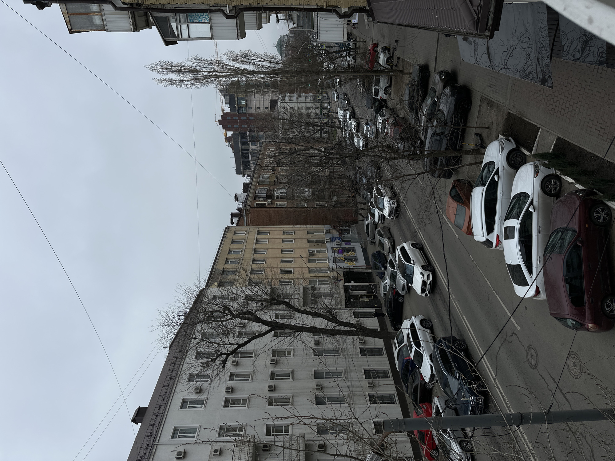 The image depicts an urban street scene in the city of Rostov-on-Don, Russia, observed from an elevated vantage point, likely a window or balcony. The sky is overcast, indicating a dull weather condition.
**Environment:**
The scene is dominated by a multi-lane asphalt road running from the foreground into the distance, bordered by sidewalks and multi-story buildings. Bare deciduous trees line both sides of the street and sidewalks, suggesting a winter or early spring season. Overhead utility wires crisscross the frame against the grey sky. A tower crane is visible in the distant background, indicating ongoing construction.
**Buildings:**
On the left side of the street, a large, multi-story building with a light-colored (off-white) facade features numerous identical windows and externally mounted air conditioning units on each floor. Its roof is dark and pitched. Adjacent to it, further down the street, is a multi-story building with a beige facade. On the right side of the image, partially visible in the foreground, is a building with reddish-brown brickwork and enclosed balconies. Various other multi-story residential and commercial buildings populate the mid-ground and background, including one under construction.
**Road and Traffic:**
The asphalt road has visible double white lines marking lanes. It is actively used by vehicular traffic and also serves as a parking area. A significant number of vehicles are parked bumper-to-bumper along both sides of the street, with additional vehicles in designated parking zones and moving in the traffic lanes. The parked vehicles primarily consist of modern sedans, SUVs, and hatchbacks, with common colors including white, black, grey, and red. Two circular manhole covers are visible on the road surface.
**People and Objects:**
One lone individual, dressed in dark clothing, is walking on the sidewalk along the left-hand building. No other specific human interactions are discernible. A small, dark-colored kick scooter or electric scooter is visible on the sidewalk near a tree on the right side of the street. Some small, green evergreen shrubs are planted in patches along the right sidewalk.