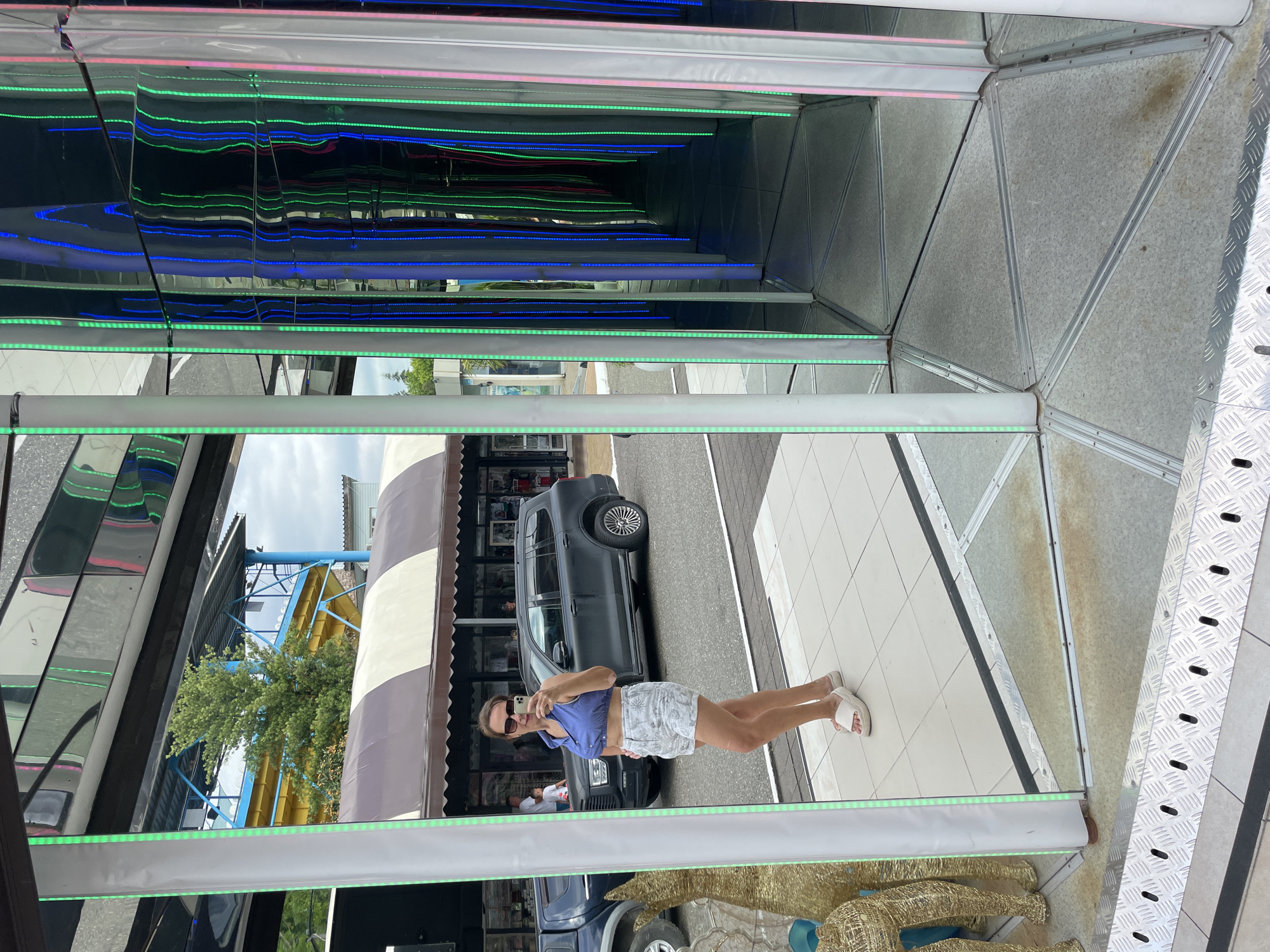 This media file shows a woman taking a selfie in a mirrored tunnel. The tunnel is decorated with colorful LED lights. The woman is wearing a blue crop top and white shorts. She is standing on a tiled floor. In the background, there is a car and some shops. The scene appears to be outdoors, possibly in a tourist area or amusement park, given the presence of a water slide in the distance. The lighting suggests it is daytime. The woman seems to be in a playful mood, as she is smiling and taking a selfie. The mirrored tunnel creates a disorienting and fun visual effect. The overall atmosphere is lighthearted and recreational. There is no visible text in the image.