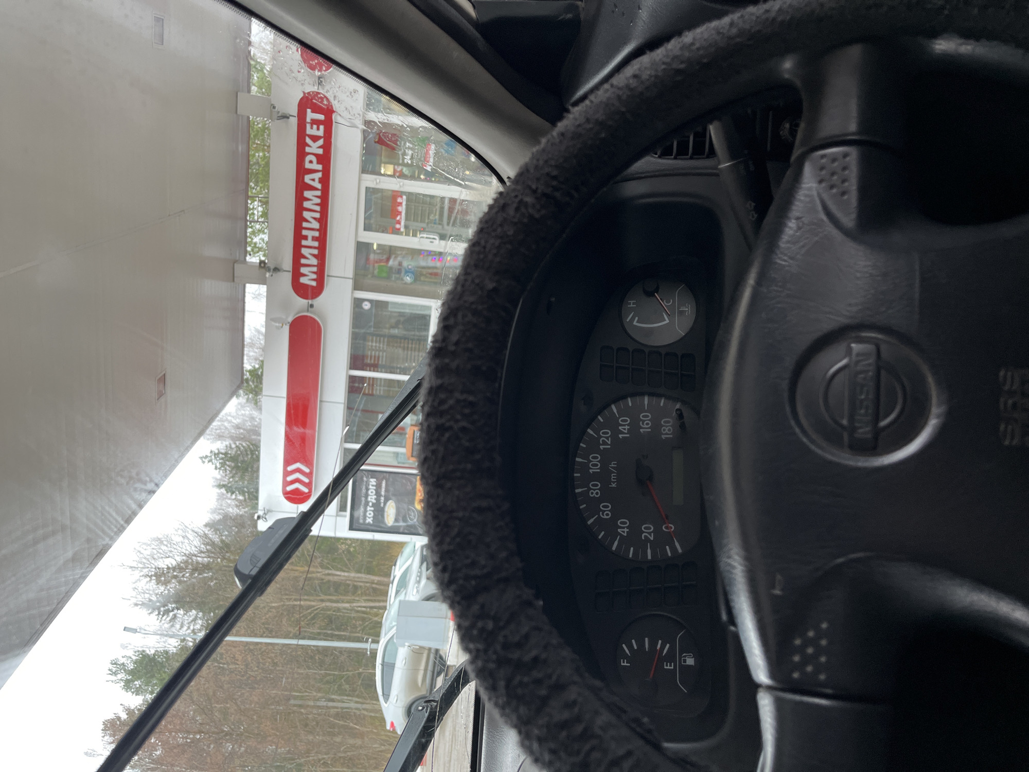 The image shows the interior of a Nissan vehicle at a "MINIMARKET" location, likely a gas station convenience store, in Chaikovsky, Russia. The dashboard includes the speedometer, fuel gauge, and temperature gauge. Through the windshield, the store's sign and exterior are visible. Several vehicles are parked outside. The environment appears overcast, and the trees have lost their leaves, suggesting it's either late fall or winter.