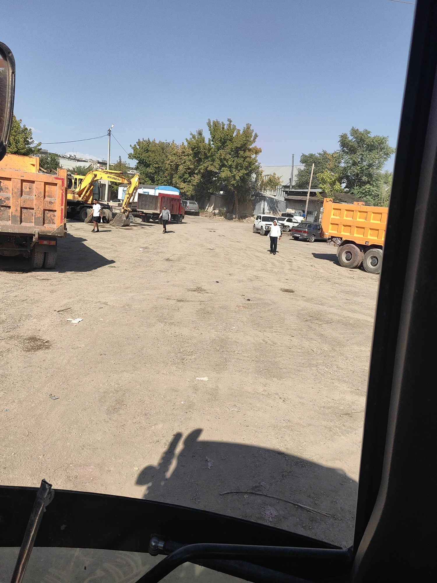 The image depicts an outdoor scene in Shymkent, Kazakhstan, likely a construction or industrial yard. A bright orange dump truck with a large, empty cargo bed is parked prominently on the left side of the frame, viewed from the rear. To its right, a yellow Hyundai excavator is positioned with its boom lowered. Several individuals are present in the scene: two men are standing near the excavator and a red trailer truck, and another man is standing near an orange truck with a large cargo bed, parked on the right side of the frame. Several cars and trucks are visible in the background, along with some trees and buildings. The ground is unpaved, appearing to be a mix of dirt and gravel. The sky is clear and blue. The foreground is partially obscured by what appears to be the interior of a vehicle, suggesting the image was taken from inside a cabin.