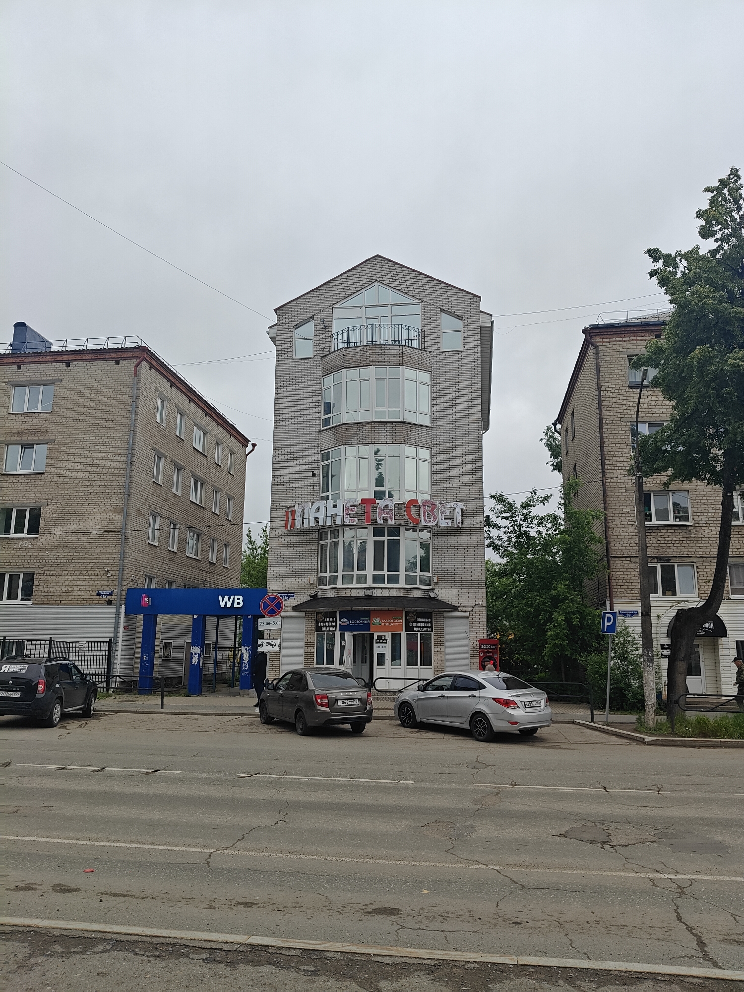 The image depicts a street scene in what appears to be the city of Chaikovsky, Russia, under an overcast sky. A three-story brick building with a distinctive architectural style stands prominently, with the Russian words "ПЛАНЕТА СВЕТ" (Planet Light) displayed on its facade. Below the building, there are two parked cars, a grey Lada and a silver Hyundai. In the background, flanking the central building, are two additional brick buildings of a simpler design.
To the left of the central building is a blue "WB" (likely referring to Wildberries, a Russian online retailer) pickup point or kiosk, and a dark-colored compact SUV is parked nearby. There is a pedestrian standing near the WB kiosk. A parking sign (marked with a blue "P") and street signs are visible to the right of the central building, near a tree with foliage. A person wearing a green military jacket is seen in the background near the tree.
The street shows signs of wear, with cracks and imperfections. There are power lines running across the top portion of the frame. The overall impression is of a typical urban scene.