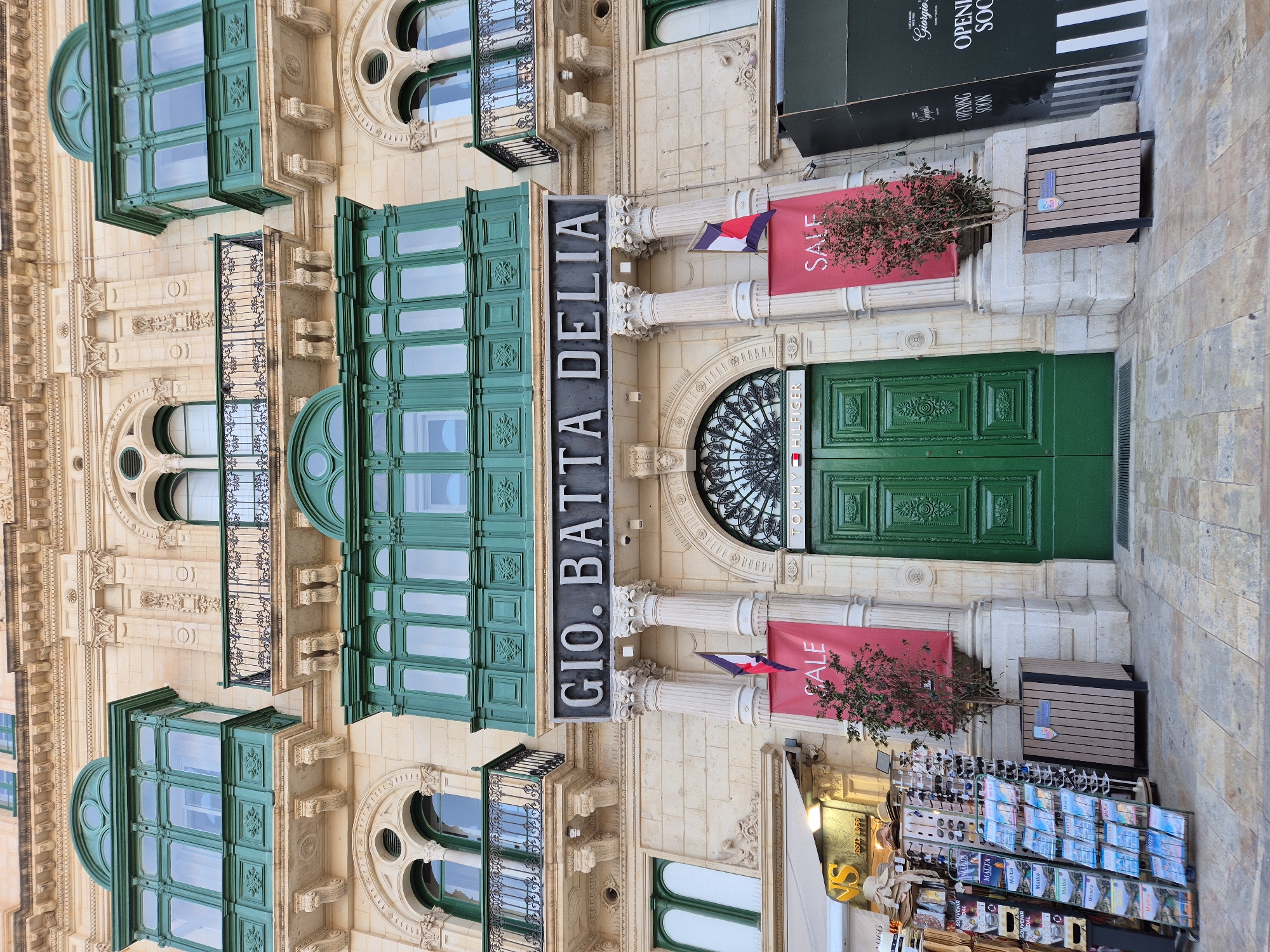 This is a low-angle, full shot of a building facade in Valletta, Malta, on a bright, overcast day. The building's architecture is ornate, with a limestone exterior and decorative elements such as columns, arches, and balconies. The most prominent feature is a large, green, enclosed balcony with multiple windows. Above this balcony, a dark sign reads "GIO. BATTA DELIA" in white lettering. Below the sign, two large, red banners with the word "SALE" are hung, one on each side of a tall, green, double door entrance. Two columns flank the entrance, and a small tree is planted in a wooden planter between them. To the left of the entrance, there is a rack of souvenirs and brochures, and a white awning above a storefront. To the right of the entrance, a large, black construction barrier with white stripes and text that says "OPENING SOON" is visible. The pavement in front of the building is made of stone. No people are visible in the image.