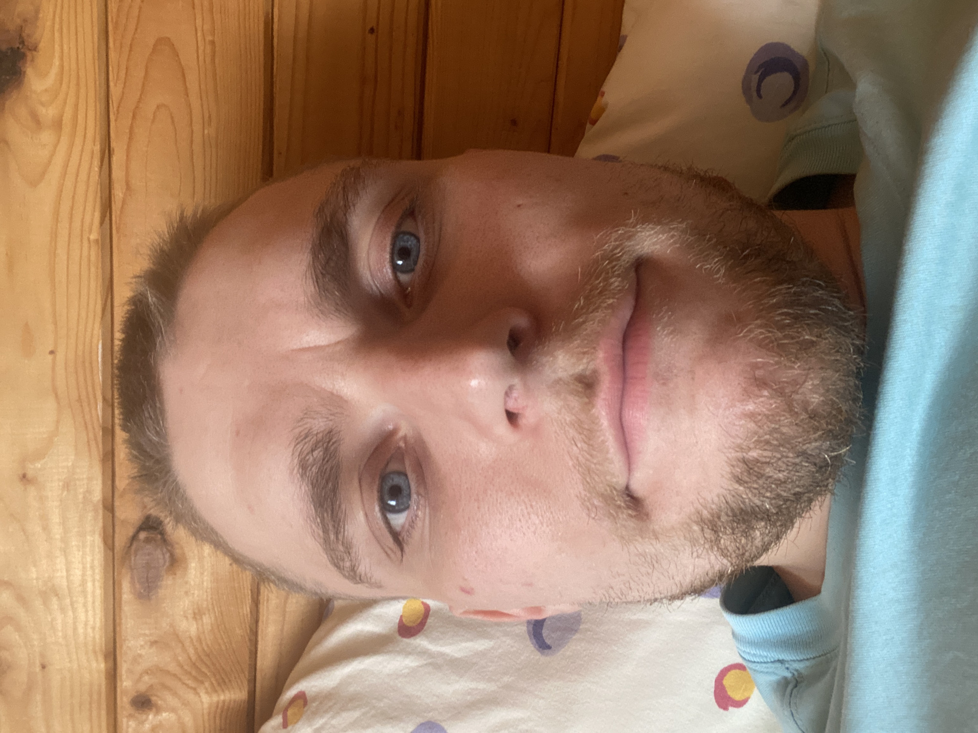 A close-up, indoor photograph shows a fair-skinned man with blue eyes and a light brown beard and mustache. He is lying down with his head resting on a pillow that has a pattern of colored circles. He is wearing a light blue shirt. The background includes a wooden wall. The image does not provide information about the city of Прикамский, Russia.
