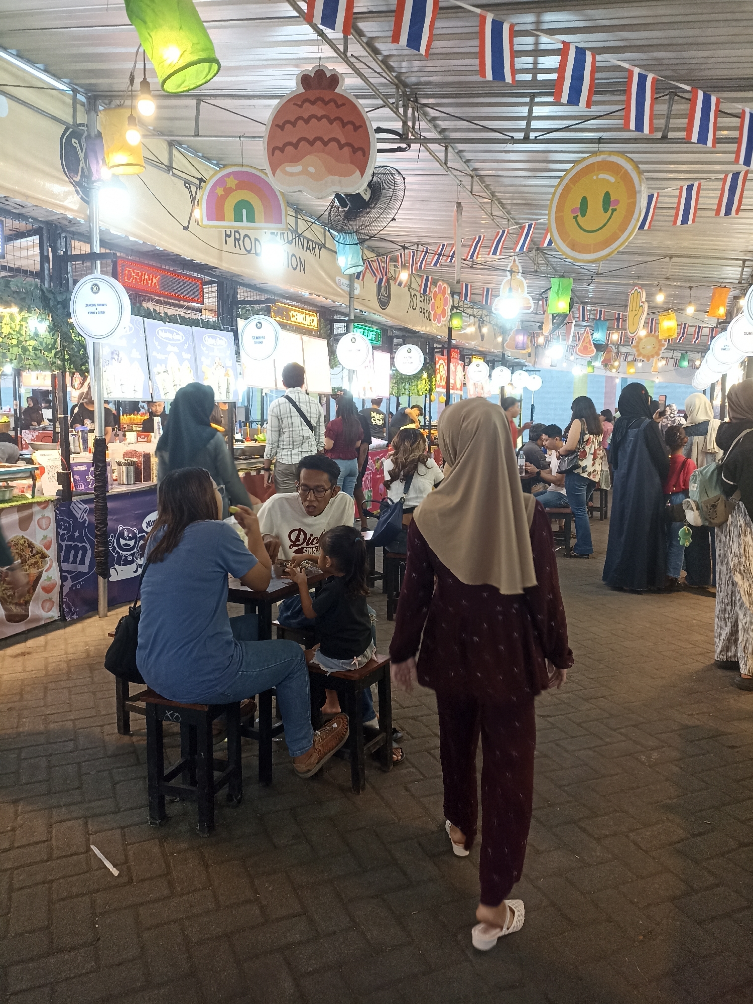 In Labruk Lor, Indonesia, an outdoor night market scene unfolds. A paved walkway is lined with food stalls, illuminated by overhead lights and decorated with strings of Thai flags and colorful paper lanterns. Several people are visible: a family of three sits at a table eating, while others stand or walk through the market. A woman in a maroon outfit and headscarf walks away from the camera, her white sandals visible. In the background, stalls display menus and food items. The atmosphere suggests a lively evening gathering for dining and socializing.