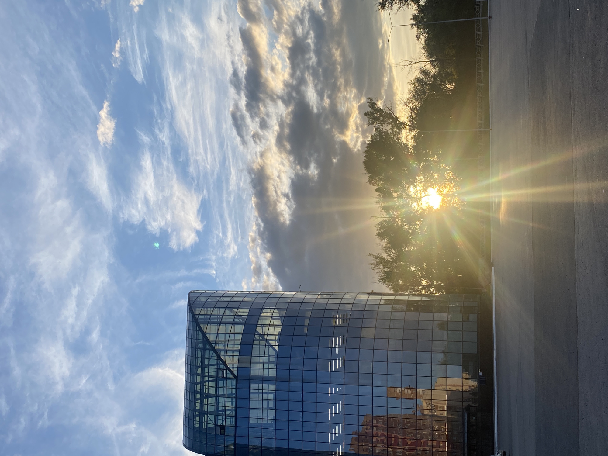 An exterior view of a modern, multi-story glass building in Samara, Russia, is captured at sunset. The building, with its reflective blue glass facade, stands to the left of the frame, showcasing rows of illuminated interior lights. In the background, a dense line of trees is silhouetted against the vibrant sky. The setting sun, positioned behind the trees, casts strong rays of light that create a lens flare effect across the lower half of the image. The sky above is a dramatic mix of blue and scattered clouds, with wispy cirrus clouds and darker, heavier formations. The ground in the foreground is an asphalt-covered area, appearing to be a parking lot or open space, with a white line marking the edge of the asphalt near the building. There are no people or vehicles clearly visible in the frame, aside from a faint outline of a white car on the far left. The overall scene depicts an urban environment at the end of the day, characterized by architectural elements and natural light.