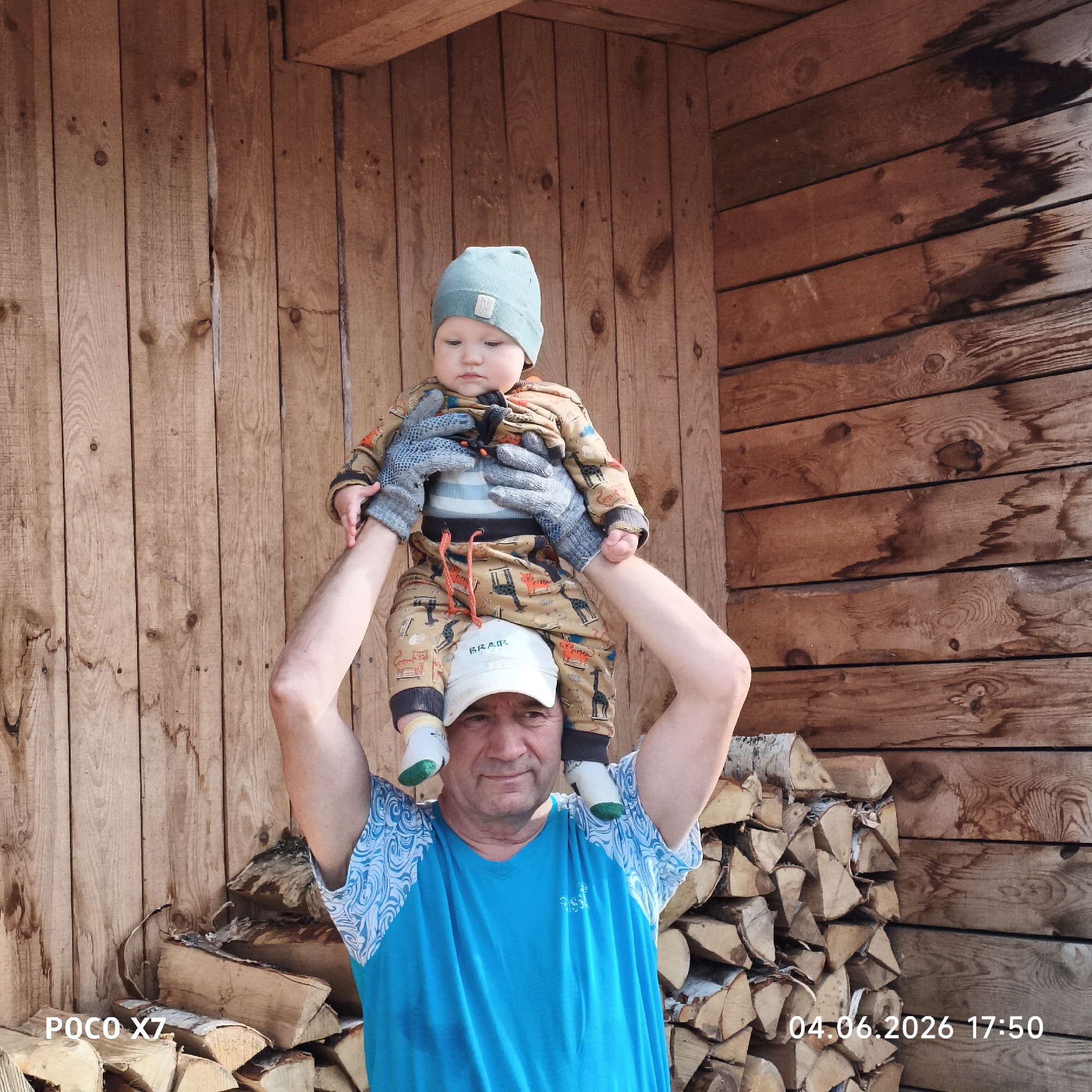 An outdoor, eye-level shot captures a man carrying a baby on his shoulders against a rustic wooden background in Chaikovsky, Russia. The man, likely a grandfather, wears a blue short-sleeved t-shirt with white patterned sleeves and a white baseball cap with "BRAIR" inscribed on it. He has a calm and slightly smiling expression as he supports the baby with his hands.
The baby, dressed in a full-body patterned outfit featuring animal designs, a light blue knit hat, and grey knitted gloves, sits securely on the man's shoulders, looking forward with a curious gaze. Their heads are centered in the frame, with the man's arms extended upwards to hold the baby's legs.
The background consists of a weathered wooden wall made of horizontal planks, likely part of a cabin or shed. To the right and bottom of the frame, a neatly stacked pile of cut firewood is visible, suggesting an outdoor, possibly rural, setting. The lighting indicates a bright, sunny daytime, casting even light on the subjects. Visible text includes "POCO X7" in the bottom left corner and a timestamp "04.06.2026 17:50" in the bottom right corner. The scene evokes a feeling of familial bond and outdoor activity.