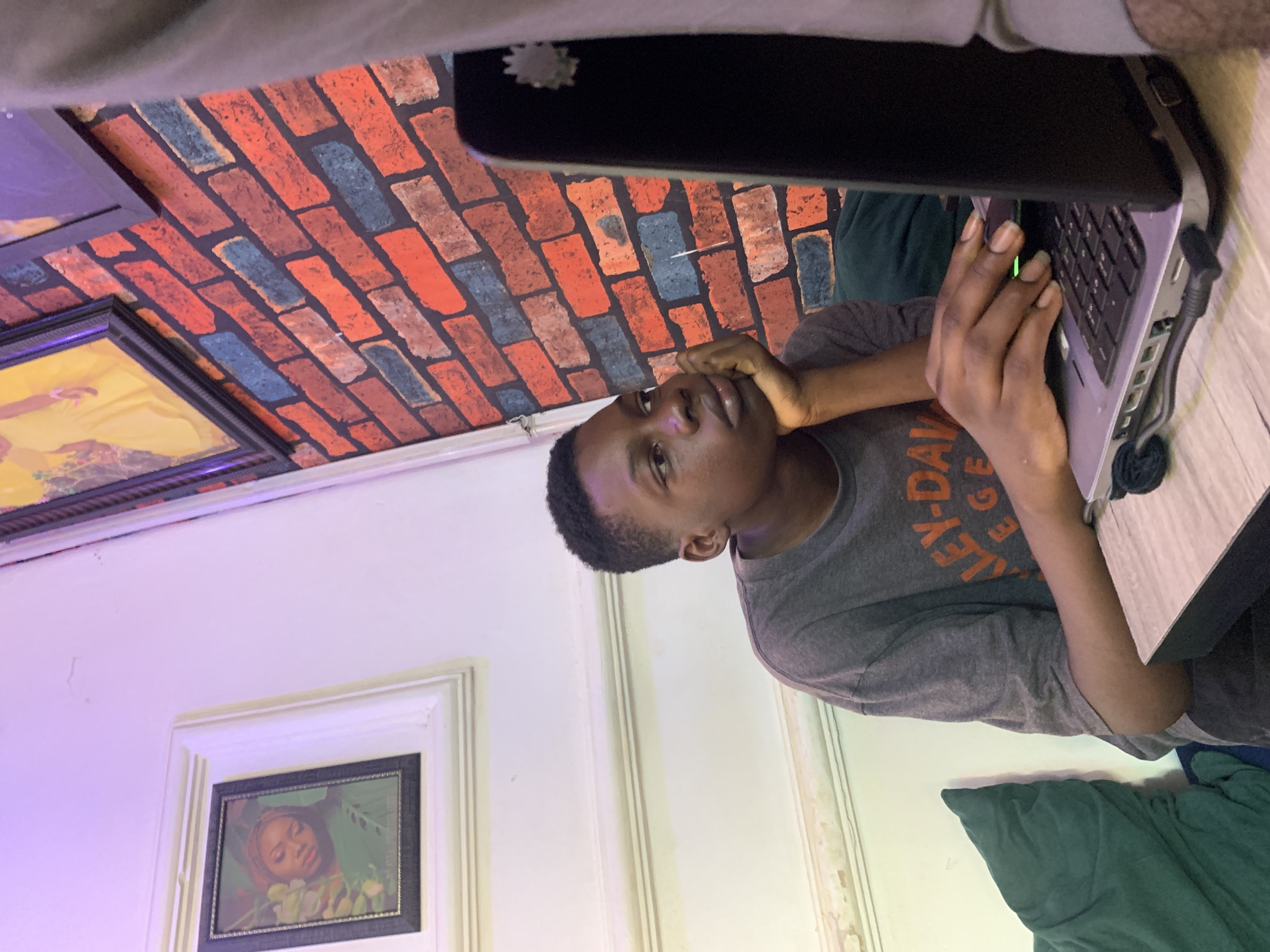 The image depicts an indoor scene in Sabojibi, Nigeria, featuring a young, male-presenting individual engrossed in an activity with a laptop. The person, with short, dark hair, is wearing a grey t-shirt that displays the text "HARLEY-DAVIDSON LEGENDARY" on its front. They are seated at a surface, looking upwards and to their right with a thoughtful or contemplative expression. Their left hand is gently resting on their chin, while their right hand is positioned over the keyboard of an open, light-colored laptop. A small, bright green light can be seen near the laptop's keyboard, indicating it is active.
The background wall behind the person features a distinctive brick-patterned wallpaper or mural, with bricks in shades of red, orange, brown, and some blue, set against a dark background. To the left, a plain white wall is visible, brightly illuminated by a strong purple light source emanating from the left side, and a subtler greenish light from the right. Two framed pictures adorn this white wall: one larger frame in the foreground contains a portrait of a person, likely a woman, surrounded by green foliage, while another yellow-toned framed picture is partially visible further up. A green or teal-colored pillow or blanket is seen on a surface to the right of the individual. The artificial lighting suggests the scene could be taking place during the evening or night. The overall atmosphere conveys a sense of quiet focus or contemplation.