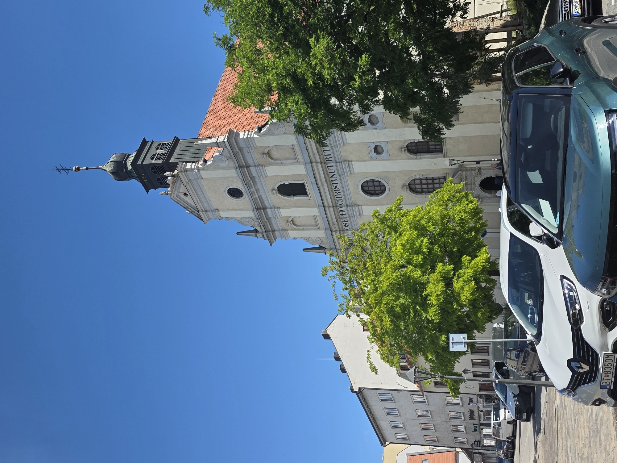 The image depicts a street scene in the city of Trnava, Slovakia, on a clear, sunny day. In the background, a light-colored church with a red tile roof and a dark green steeple is visible, adorned with a cross. The steeple has the inscription "1856". The facade of the church features architectural details and Latin inscriptions. To the left of the church, a row of white and light gray buildings with multiple windows lines the street. In the foreground, several cars are parked. A white Renault is prominently featured in the center, with a license plate showing "BT 835DM". To its right, a teal-colored SUV is partially visible. A dark sedan is parked behind the white Renault. The street appears to be paved with cobblestones or similar material. Lush green trees are interspersed throughout the scene, with one large tree in the mid-ground and another on the right side of the image. A street lamp is visible to the left of the white Renault. Two individuals are standing near the entrance of the church in the background. The sky is a bright, clear blue.