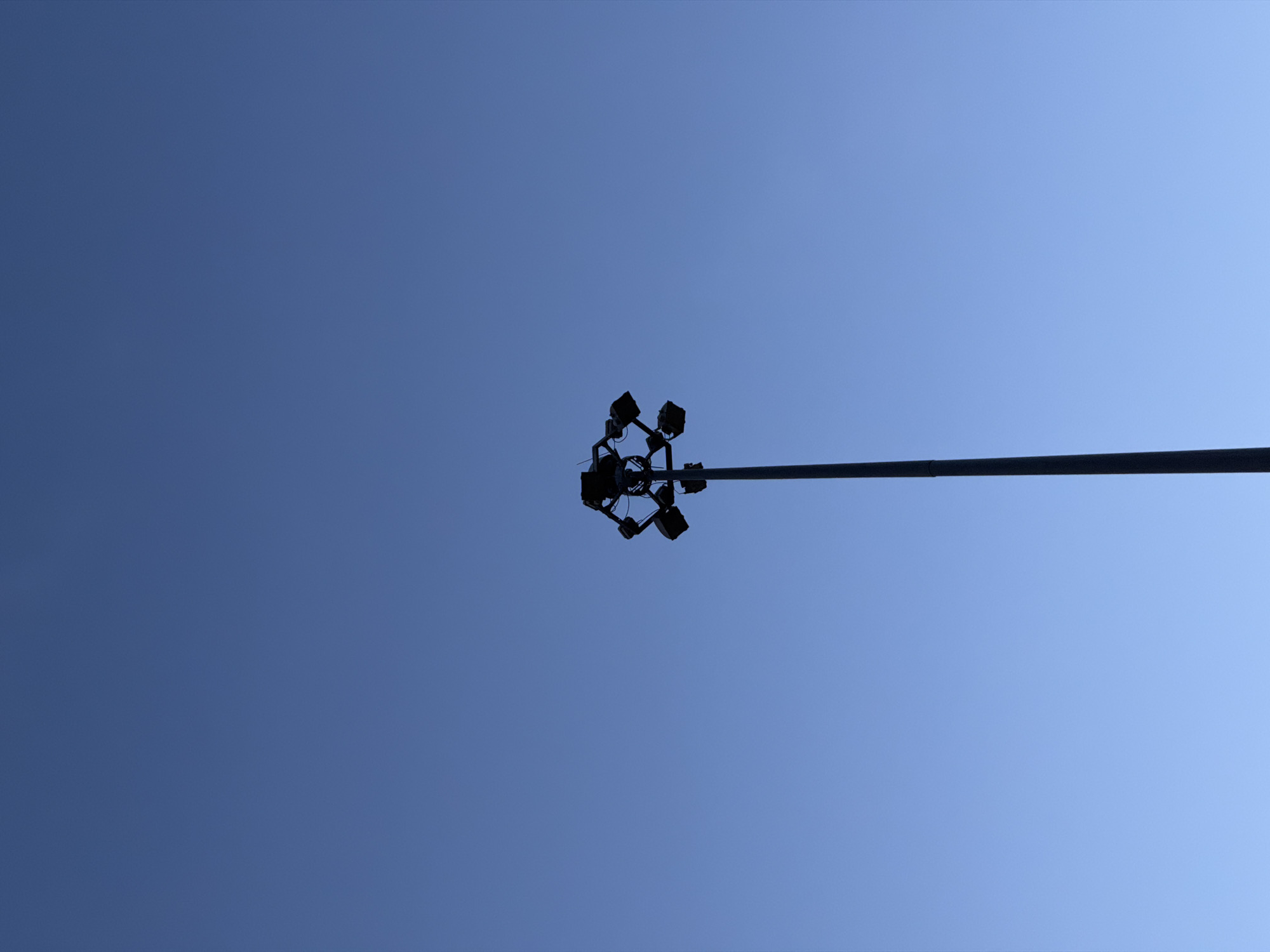 The image shows a close-up, low-angle view of a street light against a clear blue sky. The street light is a tall pole with a cluster of four spotlights at the top, all silhouetted against the bright sky. The spotlights are arranged in a circular pattern, pointing outwards. A long, horizontal arm extends from the pole, supporting the spotlights. The sky is a uniform, bright blue, suggesting it is daytime. There are no visible clouds, indicating clear weather. The setting appears to be an outdoor urban environment, likely in Krasnogorsk, Russia, given the context. No people, text, or specific events are depicted in the image. The focus is solely on the street light and the sky.
