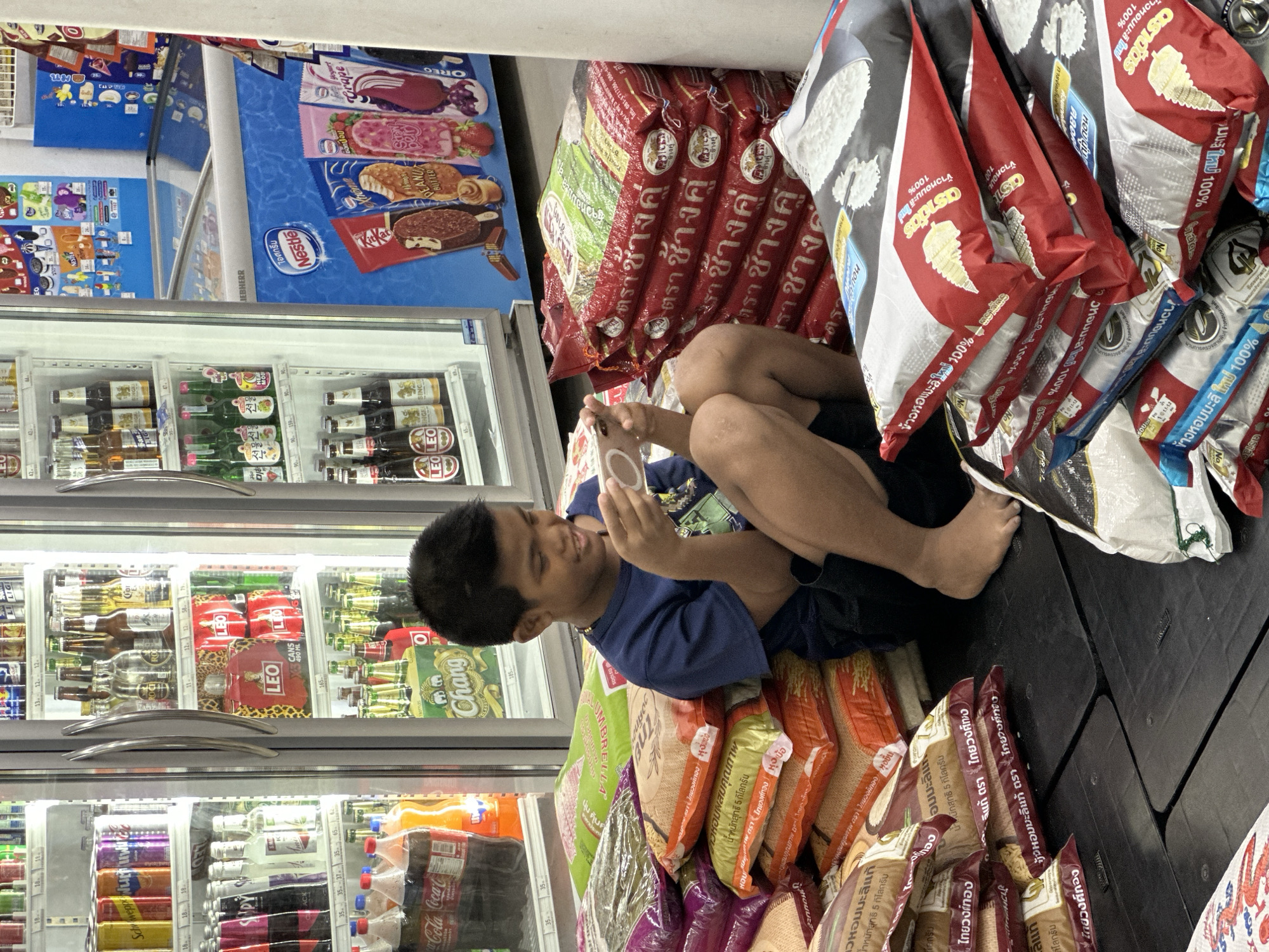 A boy is sitting on stacked bags of rice and leaning against a display cooler in a store in ตำบลเกาะพะงัน, Thailand. The cooler contains bottled beverages, cans, and ice cream treats. The boy is barefoot and wearing a blue shirt and dark shorts. He is holding a mobile phone and looking at it. The bags of rice are stacked on the floor and the cooler is stocked with drinks. There are also bags of chips stacked next to the rice.