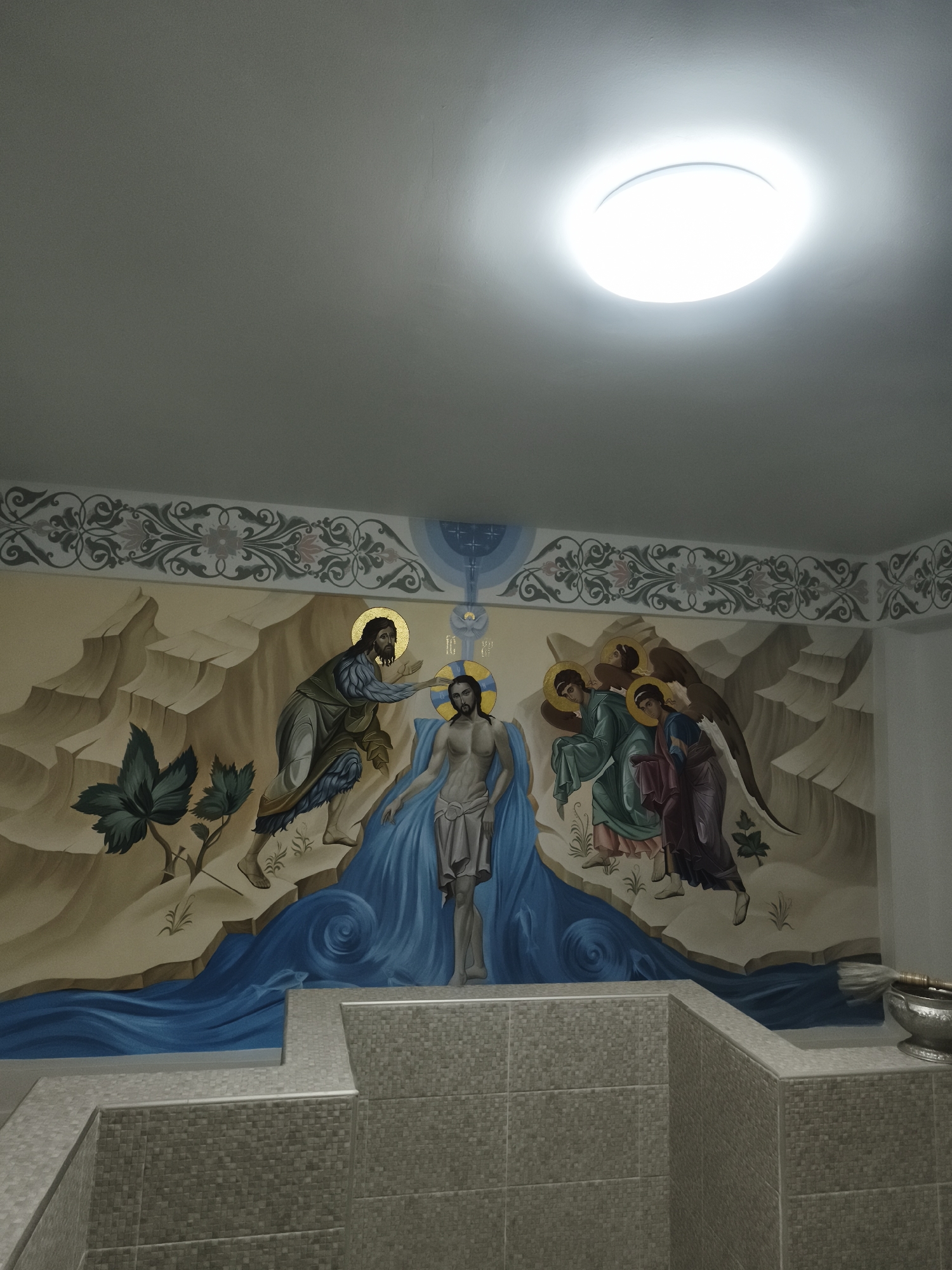 This image depicts the interior of an Eastern Orthodox baptistery located in Ko Samui, Thailand. A large, circular flush-mount LED light fixture, emitting bright white light, is centered on the light grey ceiling in the upper portion of the frame. Directly beneath the ceiling, a decorative frieze with grey floral and vine motifs runs horizontally across the wall. Below this frieze, a large mural renders the Baptism of Christ in an iconic style.
The central figure in the mural is Jesus Christ, depicted semi-nude with a white loincloth and a golden halo, standing in a stylized river of flowing blue water. To Christ's left, John the Baptist, wearing a coarse, fur-like garment and also bearing a golden halo, extends his right hand towards Christ's head. To Christ's right, three angels with golden halos and visible wings stand, appearing to hold garments or towels. Above Christ, a celestial representation features a blue arch with a golden ray and a white dove, symbolizing the Holy Spirit. The landscape background of the mural consists of stylized, angular tan and brown rocky mountains with sparse green foliage.
In the foreground, a substantial L-shaped structure, constructed from small, square light grey mosaic tiles, occupies the lower part of the scene. This structure functions as a baptismal font or pool. On the far right edge of this tiled structure, a metallic basin or bowl containing a brush-like implement, likely an aspergilium for sprinkling holy water, is present.
