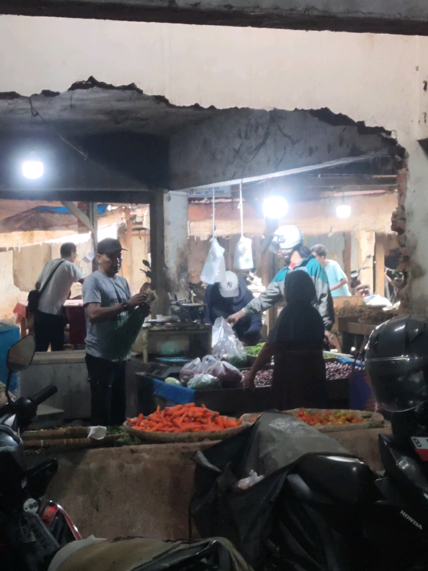A group of people are engaged in activity at a busy market stall in Karawang, Indonesia. In the foreground, a motorcycle is partially visible, covered with a black tarp. To the right, a large pile of orange carrots is prominently displayed in a woven basket. Behind the carrots, more vegetables, including what appear to be red onions, are visible.
Several individuals are interacting at the stall. A man in a gray t-shirt and black pants is holding a green plastic bag, possibly filled with produce, and appears to be weighing or preparing it. Another person, wearing a blue and gray jacket and a white helmet, is facing away from the camera, interacting with someone behind the stall. To the left, a person in a white t-shirt and black pants with a black bag slung over their shoulder is walking away. Further back, more people are visible, and the market environment suggests an active commercial area, likely outdoors or in an open-air structure, with an overhead roof and exposed ceiling elements, some of which appear damaged or incomplete. Lighting fixtures are visible, casting illumination on the scene.