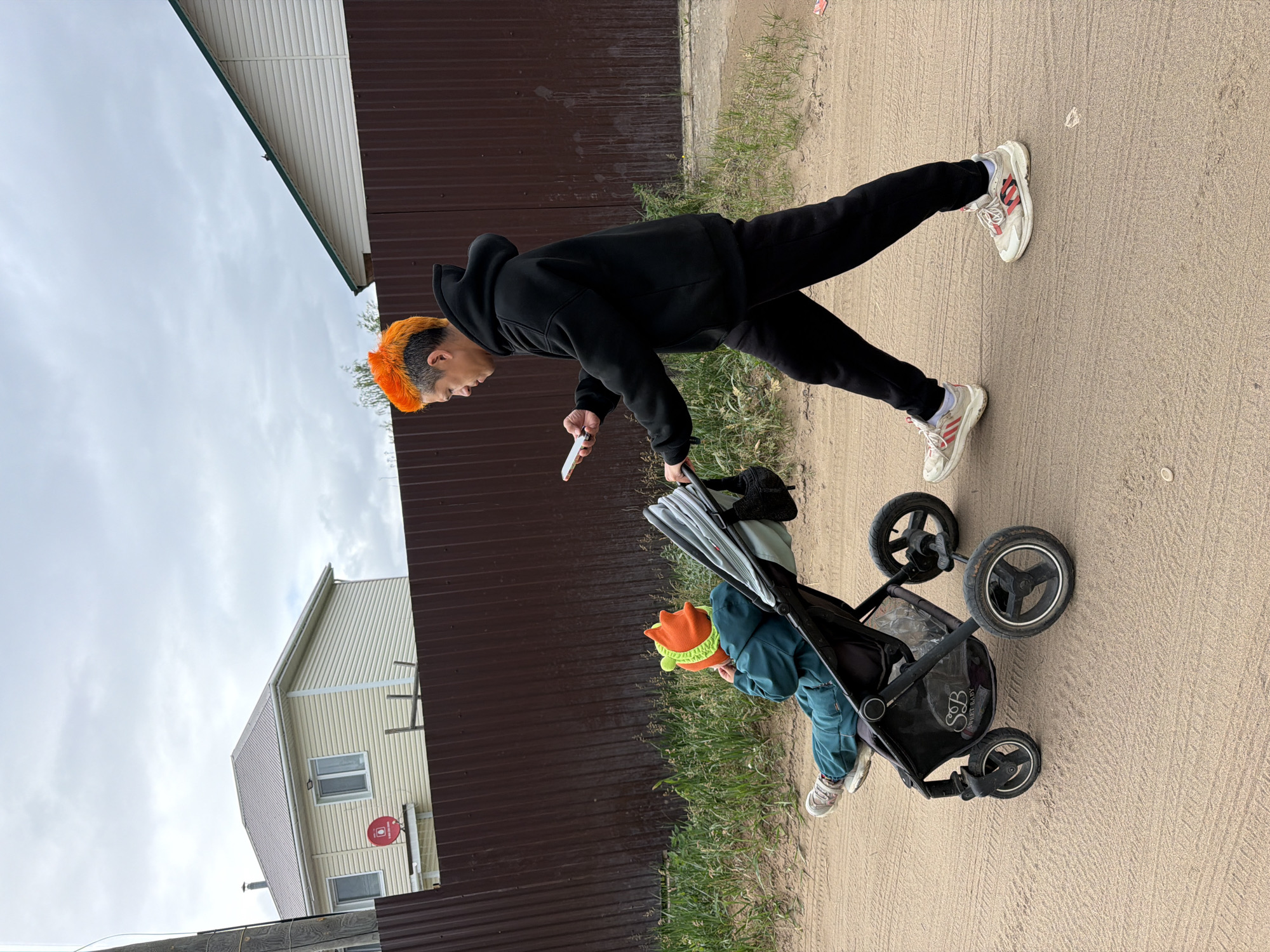 The image depicts an outdoor scene on an unpaved, sandy road in Chaikovsky, Russia, under an overcast sky. An adult with an orange and silver mohawk haircut, dressed in a black hooded sweatshirt and black sweatpants, pushes a dark-colored stroller with their right hand. The adult holds a white smartphone in their left hand and looks at its screen, walking towards the left side of the frame. White athletic shoes with red accents are visible on the adult's feet.
Seated in the stroller is a young child, facing away from the viewer and towards the left. The child wears a teal jumpsuit and an orange and lime green hat with prominent, pointed features. The stroller has at least three wheels and a storage basket beneath the seat, which shows the text "S&B SWEET BABY." A black bag hangs from the stroller's handle.
In the background, a dark brown corrugated metal fence runs horizontally across the mid-ground. Behind the fence, on the left, is a two-story house with light-colored horizontal siding, white windows, and a light brown metal roof. A red satellite dish is mounted on its side. Partially visible on the far right, behind the fence, is another house with white siding and a green-trimmed roofline. Dry grass and sparse vegetation grow along the base of the fence and at the edge of the dirt road. A wooden utility pole is visible on the far left. The sandy road shows tire tracks and ripples.