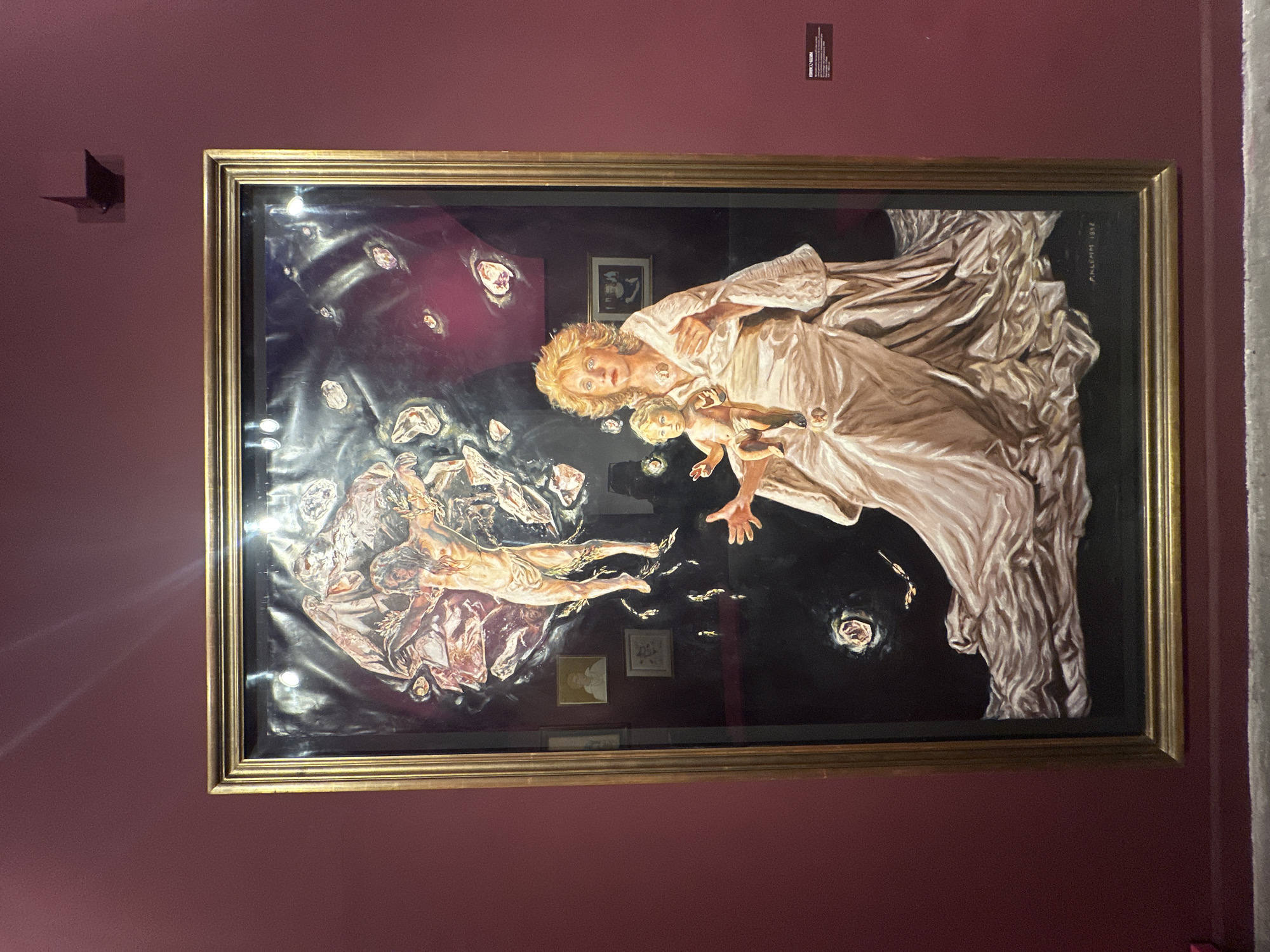 A photograph shows an artwork in a gold frame, hanging on a dark pink wall. The artwork is a painting depicting a woman in white robes holding a baby, reaching out towards a man lying on a cloud-like structure. The background is dark, with scattered objects. The painting is in the Museo Evita in the city of Buenos Aires, Argentina.