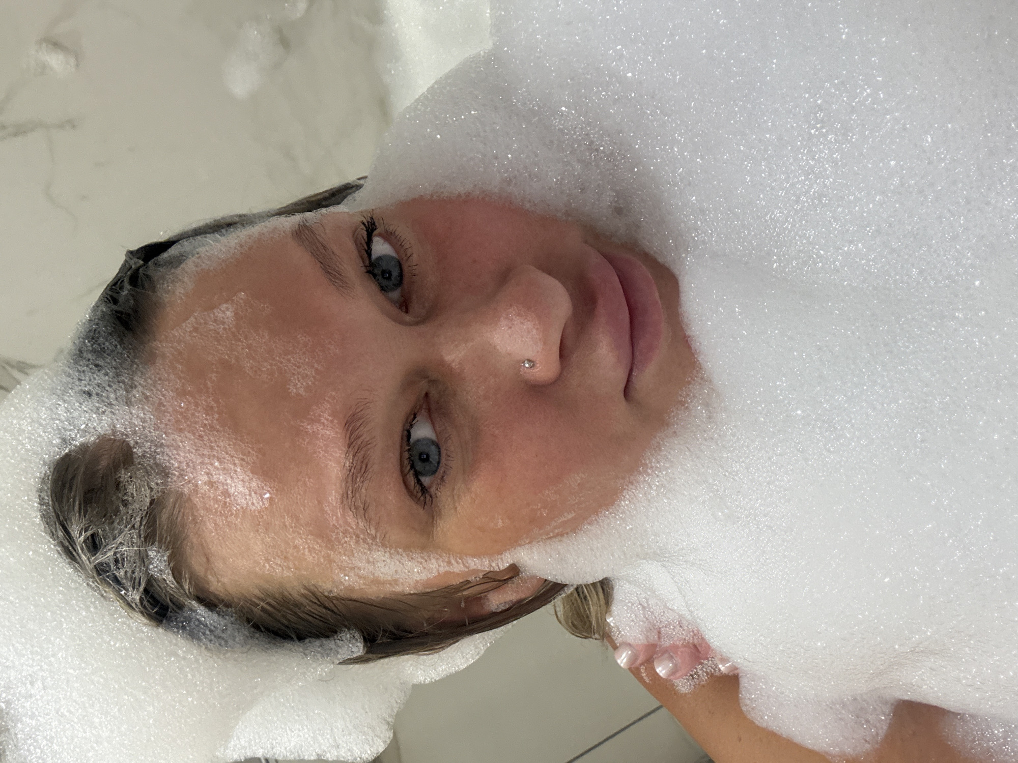 A young woman with fair skin and blue eyes is partially submerged in a bathtub filled with white foam. A small stud is visible on her nose. Wet hair is visible beneath the foam around her head. The bathroom setting appears to be modern, with light-colored tiling. The image is taken from a low angle, looking up at the woman. There is no information about the location being in Perm, Russia.