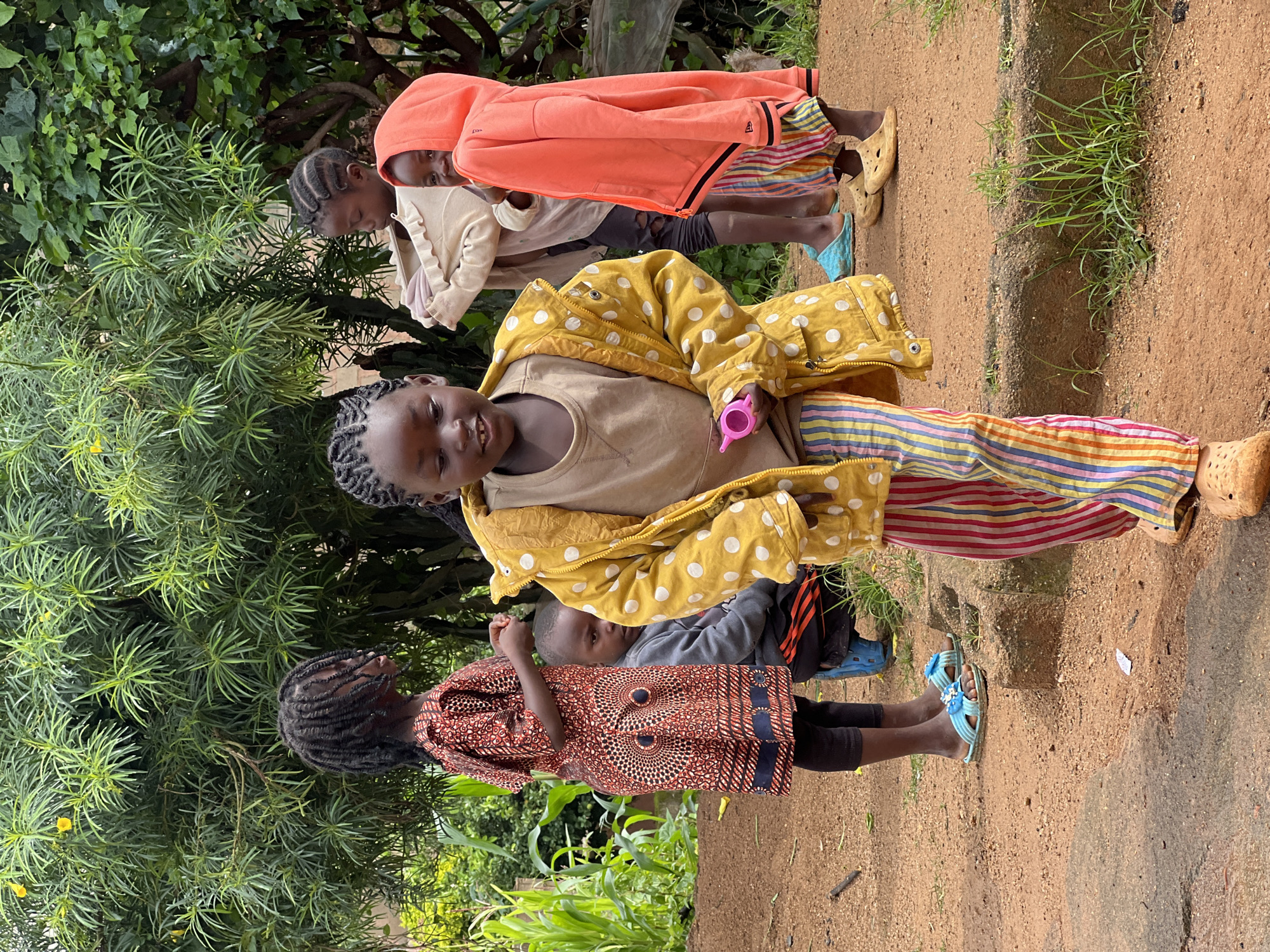 In Jos, Nigeria, a group of five young children are gathered outdoors on a dirt path, with lush green foliage and trees forming the backdrop. A young girl in the foreground, with dark braided hair, wears a bright yellow jacket adorned with white polka dots over a tan shirt and brightly colored striped pants. She holds a small pink toy in her right hand. Behind her, a girl with dark braided hair wears a red and black patterned dress and black leggings. To the far right, a young girl in an orange hooded jacket over striped pants looks towards the camera. Two other children are partially visible, one in the middle wearing a light-colored sweater and the other to the left in a grey sweatshirt. The ground is a mixture of red dirt and some patchy grass, with a low concrete border visible. The lighting suggests it is daytime, with natural light illuminating the scene.
