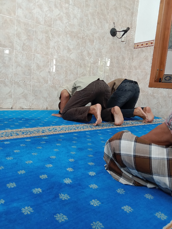 Three men are praying on a blue prayer rug with a beige and white border. They are all wearing traditional Islamic clothing. The two men in the foreground are kneeling with their foreheads on the rug. The man in the background is sitting with his legs crossed and his hands clasped in his lap. The walls are tiled and the room is lit by natural light from a window in the background. The location is a mosque in Kecamatan Brangsong, Indonesia, at coordinates (latitude: -6.948, longitude: 110.231).