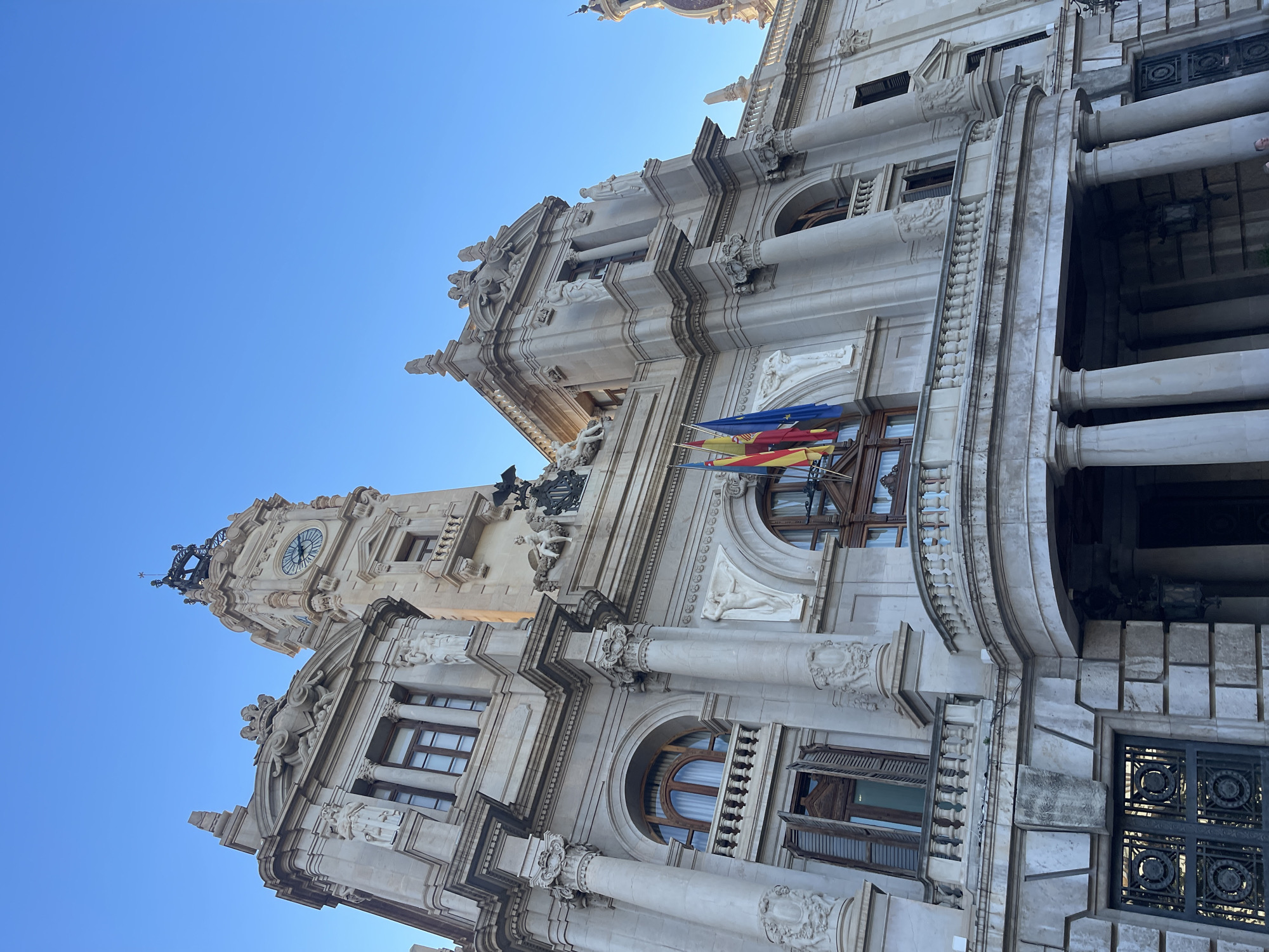 The image depicts the City Hall of València, Spain, viewed from a low angle, showing the building's facade and clock tower against a clear blue sky. Architectural details are prominent, including columns, statues, and ornate decorations. Three flags are visible hanging from a window. The facade is constructed from light-colored stone. The time visible on the clock tower face appears to be around 10:25 or 22:25. The image is vertically oriented and tilted to the right.