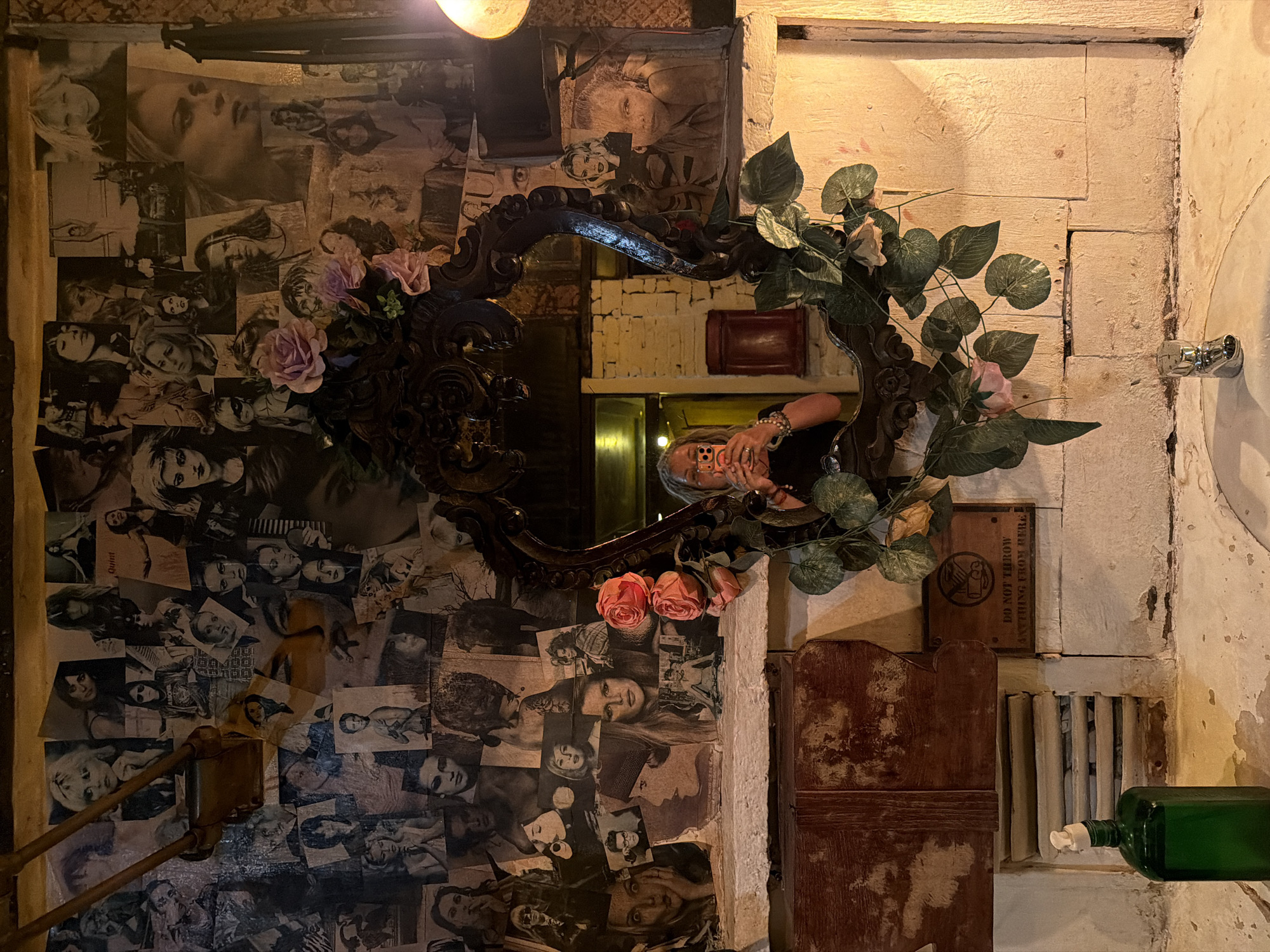 The image shows a woman taking a selfie in a mirror. The mirror is ornate and framed with artificial flowers and greenery. The wall behind the mirror is covered in a collage of black and white photographs of women. Below the mirror is a sink with a faucet and a green bottle of liquid. To the left of the sink is a wooden vent with a sign that reads "DO NOT THROW ANYTHING FROM HERE". The overall scene appears to be in a bathroom or powder room with a vintage or eclectic aesthetic. The text "DO NOT THROW ANYTHING FROM HERE" suggests the location is likely in a public or communal space, possibly a bar or establishment in Kuta, Indonesia.
