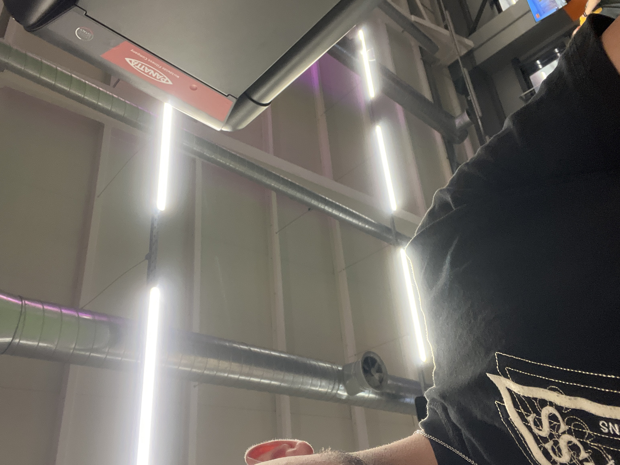 The image shows a close-up, low-angle shot from the perspective of a person in a gym, looking up towards the ceiling and to their right. The person is wearing a black t-shirt with a white graphic design. The gym ceiling features exposed metal ductwork and bright fluorescent lighting. To the right, a portion of a piece of gym equipment, possibly a treadmill, is visible with the brand name "PANATTA" clearly displayed on a red panel. The overall environment suggests an indoor fitness facility. There is no indication of the city of Plovdiv, Bulgaria, within the image itself.