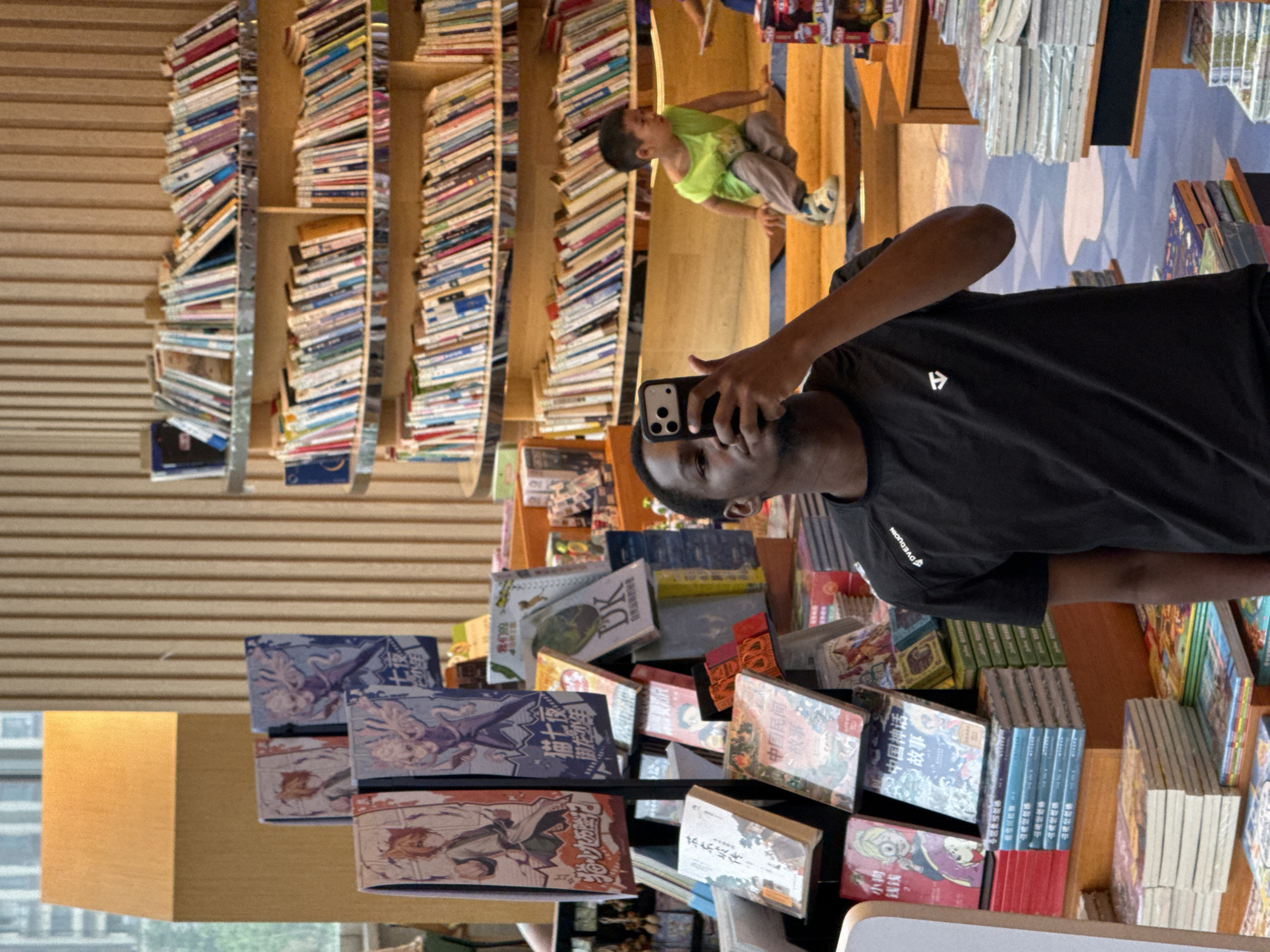 A Black man is taking a selfie in a brightly lit bookstore. He is wearing a black t-shirt with a white logo on the left chest and is holding a smartphone in his right hand. The background shows rows of bookshelves filled with books, and a young child in a green shirt is visible on a stepped platform in the distance. The man appears to be in the city of Liangjiang, China.
