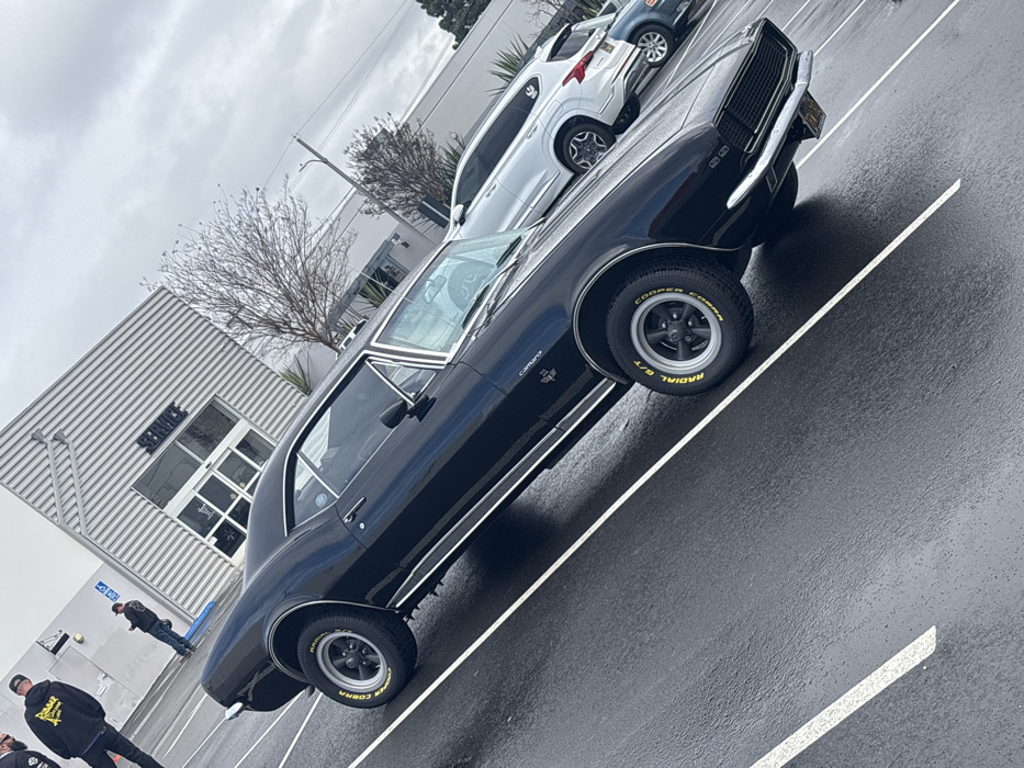 The image captures a classic black Chevrolet Camaro SS, likely a first-generation model, parked on a wet asphalt surface. The car is the primary subject, positioned diagonally across the frame, showcasing its sleek design and shiny, rain-slicked body. The "Camaro" and "SS" badges are visible on its front fender, and the tires are distinctly marked "COOPER COBRA RADIAL G/T" with yellow lettering, mounted on black multi-spoke wheels with polished rims. Chrome accents, including bumpers, window trim, and side molding, gleam against the dark paint.
The setting appears to be a parking lot outside a commercial building. The building has a modern, industrial aesthetic with light-colored corrugated metal siding and a large windowed entrance area. The word "SERVICE" is prominently displayed on the building's glass. Behind the Camaro, other vehicles are parked, including a white SUV (possibly a Toyota RAV4 or similar) and parts of another dark car. A small, bare-branched tree stands between the building and the parked cars, suggesting a winter or late autumn season.
The sky is overcast and uniformly gray, contributing to the soft, diffused lighting. The wet pavement indicates either recent rainfall or ongoing drizzle. Two individuals, dressed in dark casual wear (possibly hoodies), are partially visible in the lower-left corner of the frame, near the building. Another person is seen standing further back near the "SERVICE" building. No specific activity is clearly depicted beyond the static scene of the parked car and people standing nearby. The photograph itself is taken at a steep, upward-tilted angle, giving the impression that the ground is sloped and adding a dynamic, albeit somewhat disorienting, perspective to the scene.