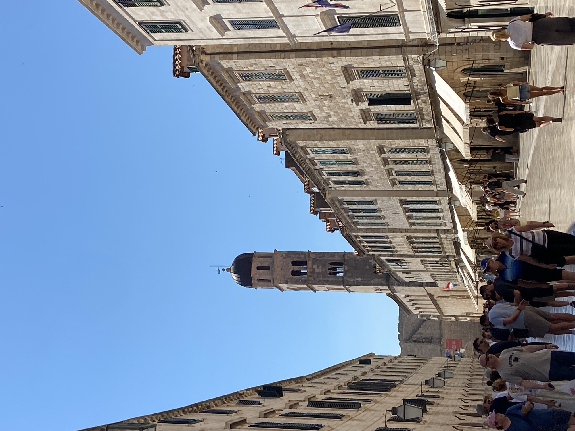 The image shows a sunlit, pedestrianized street in Dubrovnik, Croatia. The street is lined with tall, light-colored stone buildings with green shutters. A prominent bell tower with a dark dome rises in the distance. Numerous people are walking along the street, some singly, some in small groups. Many are dressed in summer clothing. Awnings extend from some of the buildings, providing shade. The sky is clear and blue. The scene appears to be a popular tourist area.