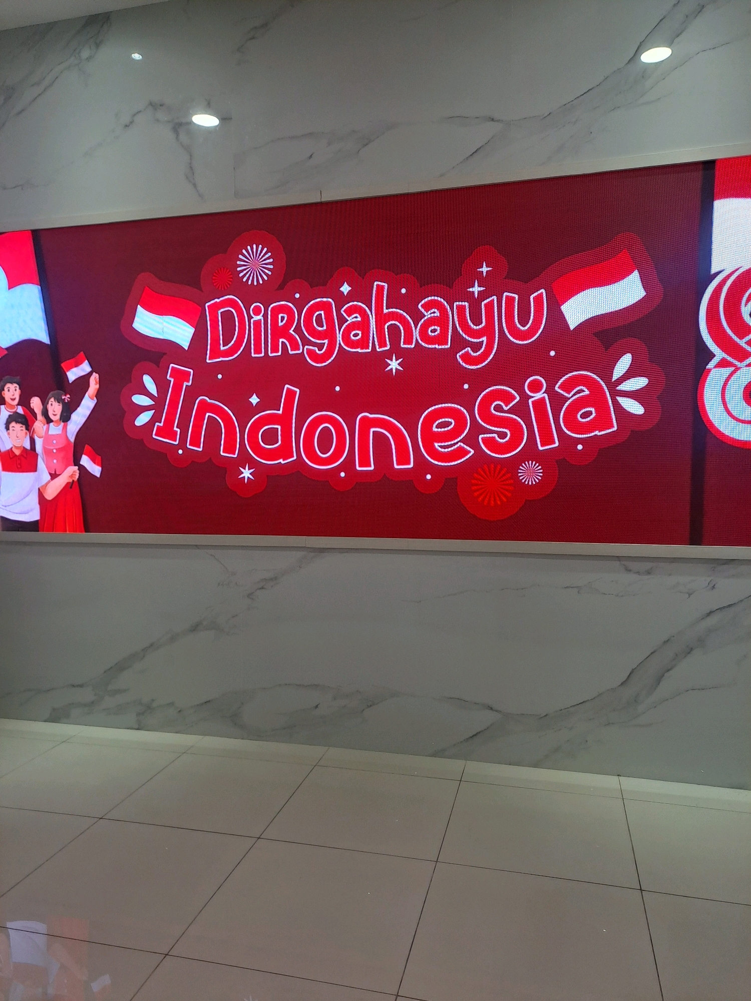The image shows a large LED screen displaying a festive message in Indonesian, "Dirgahayu Indonesia" (Happy Independence Day Indonesia), accompanied by stylized Indonesian flags and cartoon figures of people celebrating. The screen is mounted on a red background with decorative elements like fireworks and stars. To the left of the screen, cartoon figures of four young people are depicted, two of whom are holding small Indonesian flags. They are dressed in red and white attire, consistent with the national colors. The screen is positioned on a wall with a marble-like finish. Below the screen, a tiled floor is visible. The scene appears to be an indoor public space, possibly a community center or a commercial establishment, decorated for a national holiday. There are no specific landmarks or elements in the image that would allow for precise localization to a specific city like Pagerwojo, Indonesia, but the message and imagery are indicative of a celebration of Indonesian Independence Day.