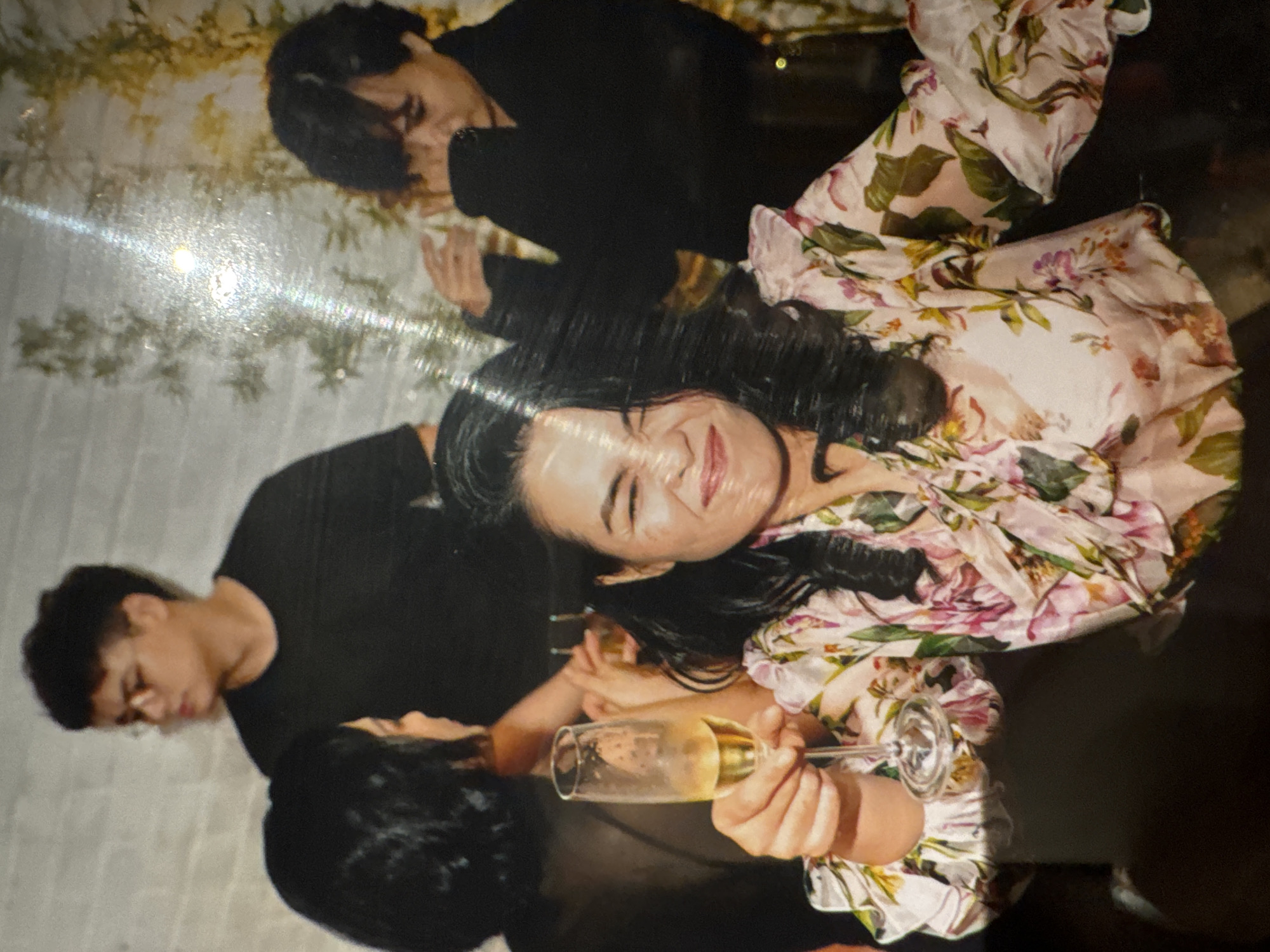 The image depicts a group of people at an indoor gathering. In the foreground, a woman with dark hair is smiling and holding up a champagne flute filled with a light-colored beverage. She is wearing a light-colored, floral-patterned top with voluminous sleeves. Behind her, a person with dark hair is partially visible on the left, and further back, a man wearing glasses and a dark t-shirt is looking to the right. On the right side of the image, another individual with dark hair is also visible. The background appears to be a wall with some decorative foliage. The lighting suggests an evening or dimly lit setting. There is no information in the image to suggest a specific location in Hà Nội, Vietnam.