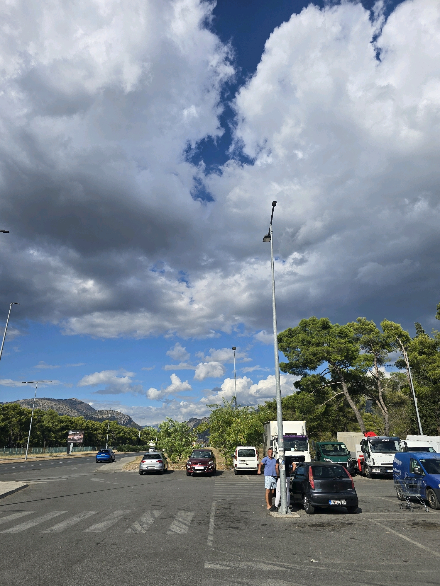 The image captures a parking lot in Podgorica, Montenegro, under a dynamic sky with both dark storm clouds and bright blue patches. In the foreground, a man in a blue shirt and checkered shorts leans against a lamppost, looking towards a black Fiat Punto parked nearby. Several other vehicles, including cars, a white van, a green truck, and two white trucks with trailers, are visible in the parking lot. The parking lot is paved with asphalt and marked with white lines, including a pedestrian crossing. Beyond the parking lot, a road runs parallel to a tree line and a billboard. In the distance, mountains form the backdrop. The overall scene suggests a daytime setting with fluctuating weather conditions.