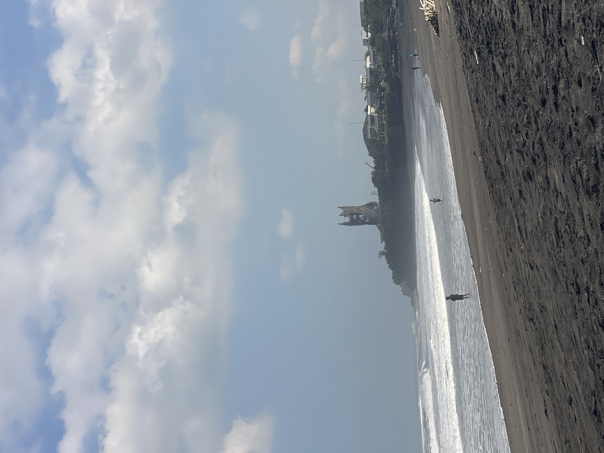 The image depicts a black sand beach with a gentle surf. In the distance, a cliff overlooks the ocean, topped with a cluster of buildings and a prominent, unusual, tower-like structure. Several individuals are visible on the beach; one stands in the shallow water, appearing to fish or wade. Further down the beach, two more figures are also engaged in activities near the water's edge. The sky is partly cloudy with patches of blue. The scene appears to be located in Kediri, Indonesia, suggested by the context provided.