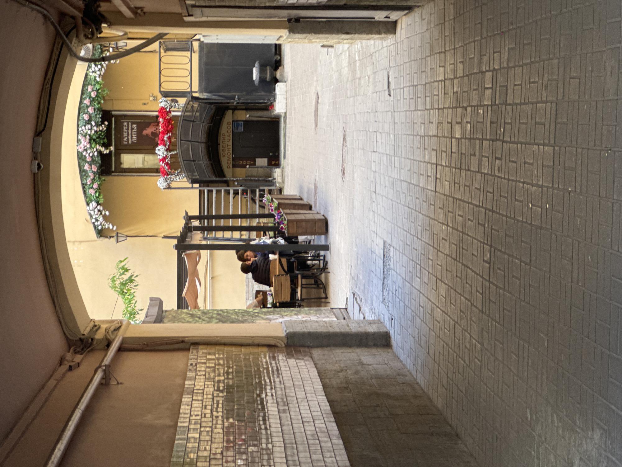 An archway frames a courtyard in Saint Petersburg, Russia, with a tiled pathway leading into it. To the left, a textured wall with rectangular tiles runs alongside the pathway. On the right, an open doorway leads into a building. Further into the courtyard, there is a seating area with a few people seated at tables. Above the entrance to the building, a balcony is adorned with flowers, and a sign reads "ГАЛЕРЕЯ ЛИТЬЯ" (Gallery of Castings). Another sign below the balcony indicates "ЮВЕЛИР РЕМОНТ ЧАСОВ" (Jeweler Watch Repair). The overall scene depicts a quaint urban courtyard with elements of commerce and leisure.