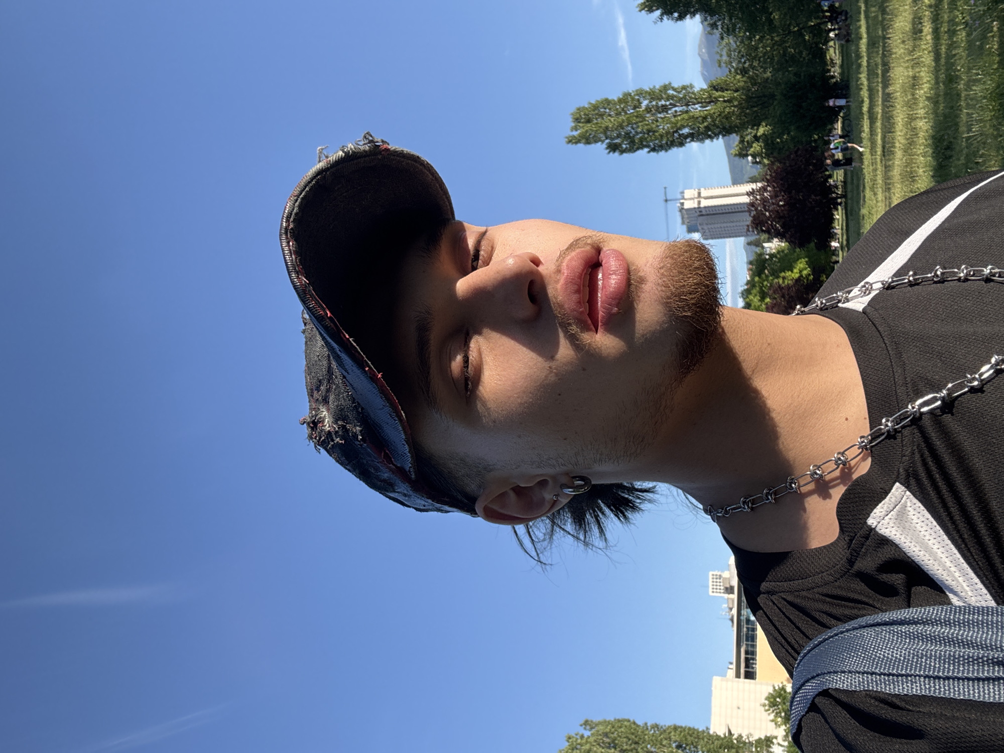A low-angle shot shows a young adult male with light skin, stubble beard, and closed eyes wearing a distressed black baseball cap. He has a silver chain necklace and a black T-shirt with white stripes. The background reveals a park with green grass and trees, under a clear blue sky. A tall building under construction is visible. In the distance, other people can be seen walking. The location is likely a park within the city of Sofia, Bulgaria.