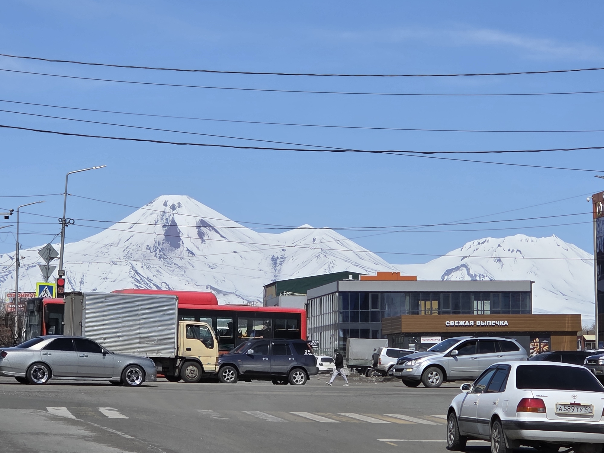 The image depicts an urban street scene in an unidentified city. In the background, snow-covered mountains dominate the skyline. Power lines stretch across the upper portion of the frame. The street is busy with vehicular traffic, including several cars, a red bus, and a light-duty truck. A pedestrian is crossing the street at a crosswalk. Buildings of modern architecture are visible, including a brown sign in Russian that reads "СВЕЖАЯ ВЫПЕЧКА" which translates to "Fresh Pastries". The weather appears sunny with a clear blue sky.