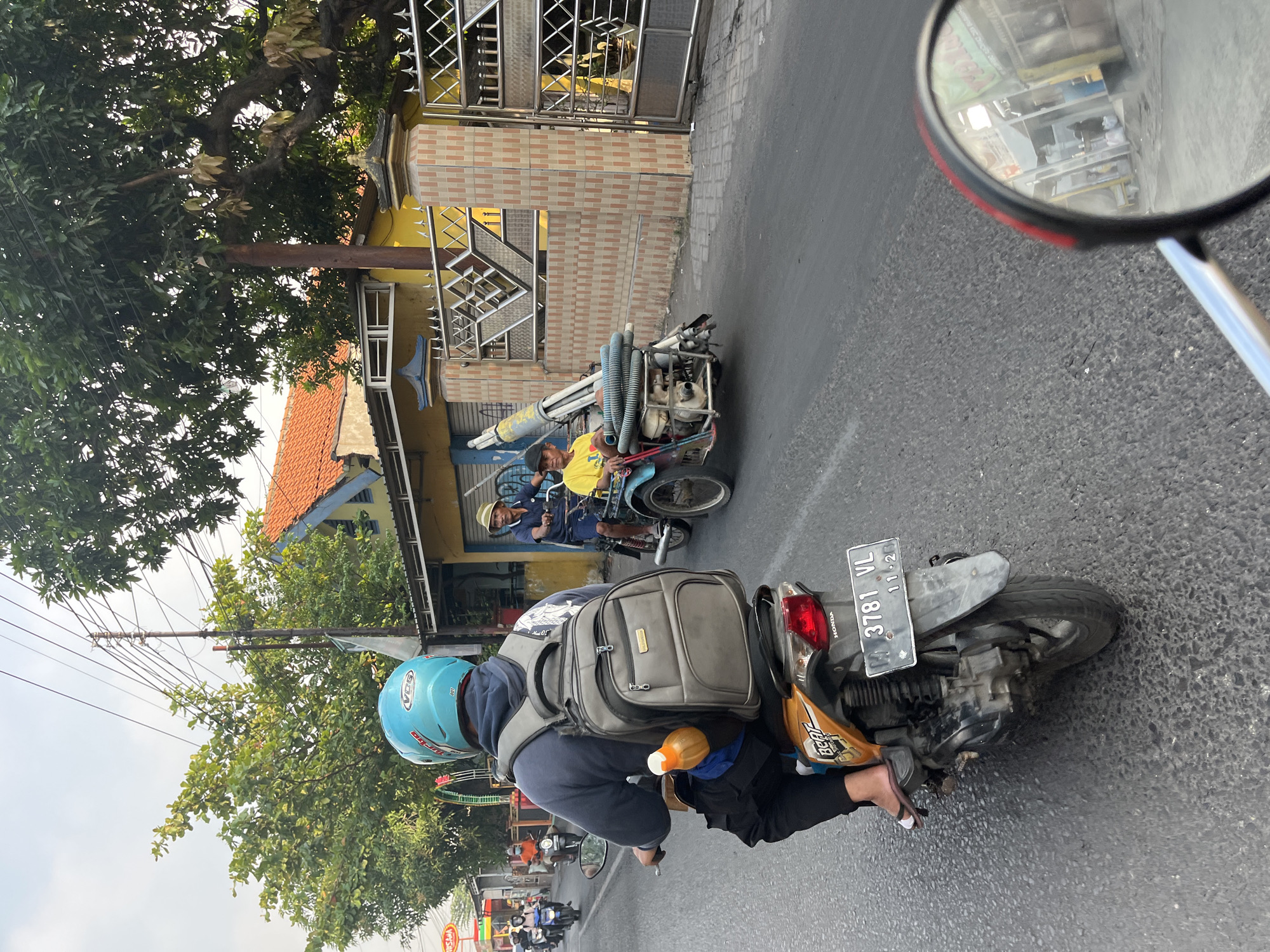 In Kedungkendo, Indonesia, a street scene unfolds with several motorcycles and a three-wheeled vehicle. The foreground is dominated by the rear view of a person riding a Honda Beat motorcycle, distinguished by its orange and black color scheme and a license plate reading "3781 VL 11-21." The rider wears a blue helmet and a grey backpack, with a yellow bottle attached to the backpack's strap. To the right, a three-wheeled vehicle, possibly a modified motorcycle with a sidecar, carries two individuals. The driver, wearing a straw hat, is looking forward, while a passenger in a yellow shirt sits beside him. This vehicle appears to be transporting a long, cylindrical object with coiled hoses. The background reveals buildings with orange tiled roofs and yellow facades, alongside trees and a metal fence. The road surface is asphalt, showing signs of wear. The overall impression is a snapshot of daily life and transportation in this Indonesian locale.