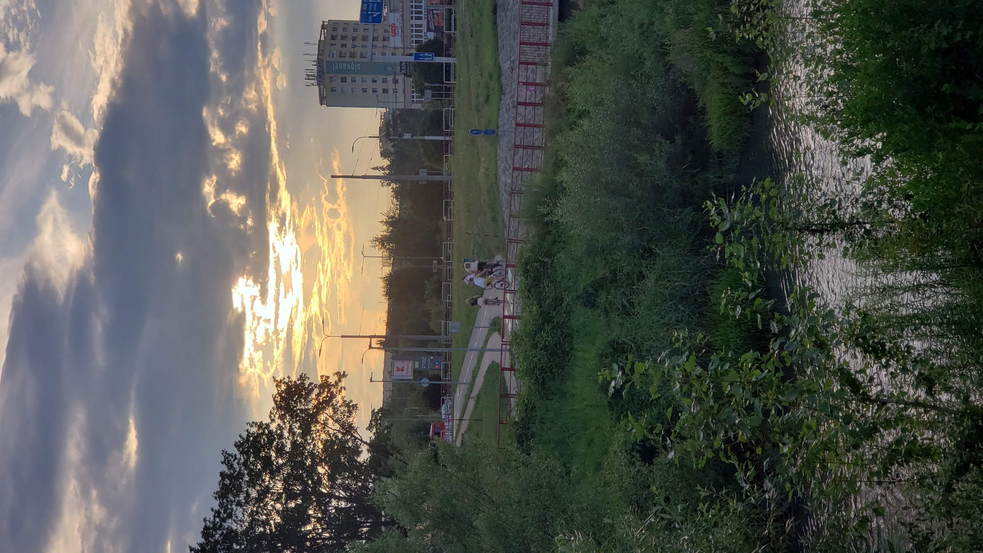This is a vertical, outdoor, eye-level photograph taken in Prešov, Slovakia. The scene captures a portion of a river in the foreground, bordered by dense green foliage. Beyond the foliage, a paved pathway runs alongside a red-railed bridge. Several individuals are seen cycling on the pathway, their forms silhouetted against the backdrop of a building and the sky. The sky is filled with dramatic clouds, with the sun breaking through in a bright, golden glow. A tall, multi-story building, likely an apartment complex or office building, is visible in the mid-ground, and further in the distance, trees and other structures indicate an urban or suburban setting. The overall lighting suggests either dawn or dusk, with the sun's rays creating a warm, diffused light across the scene. The photograph appears to have been taken from a slightly elevated position, looking across the river and pathway towards the building and sky.