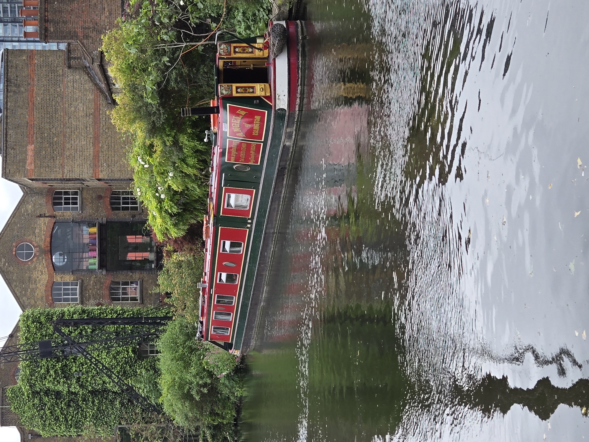 A long canal boat is moored alongside a brick building with foliage in London, United Kingdom. The boat is painted in red and green, with a sign that reads "ANGEL of ISLINGTON". The brick building has a crane covered in ivy. The water reflects the boat and the surrounding foliage.