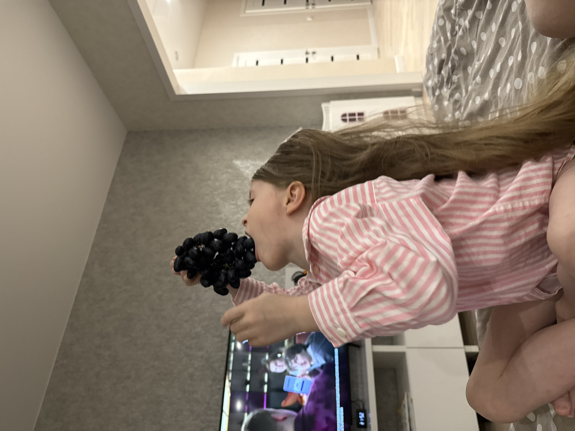 A child is eating a bunch of grapes in a room with grey walls. The child is wearing a pink and white striped shirt and is kneeling. A television is on in the background, displaying an image of animated characters. A shelf is located beside the television. A bed with a grey and white patterned comforter is visible. The image shows an upper area of the interior of the house. There is no indication of the location being Izhevsk, Russia.