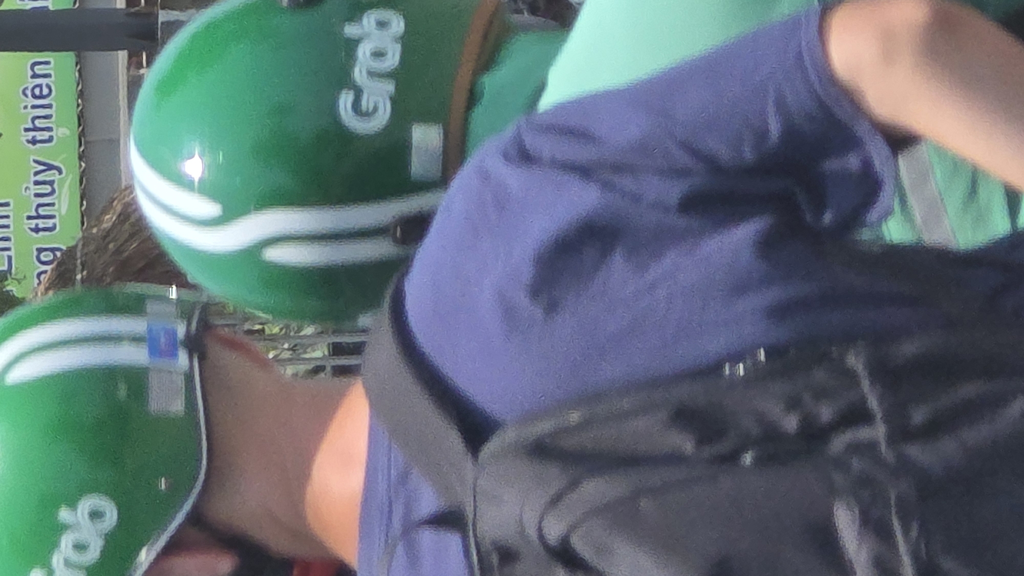 Two individuals are visible in the image, both wearing green motorcycle helmets branded with the word "Grab," indicating they are likely operating ride-hailing services. The visible individual is wearing a navy blue shirt and has a black backpack strapped on. A sign with Vietnamese script is also visible in the background, indicating the location is likely in Vietnam, specifically Nha Trang. The background is blurred, making it difficult to identify the specific location.