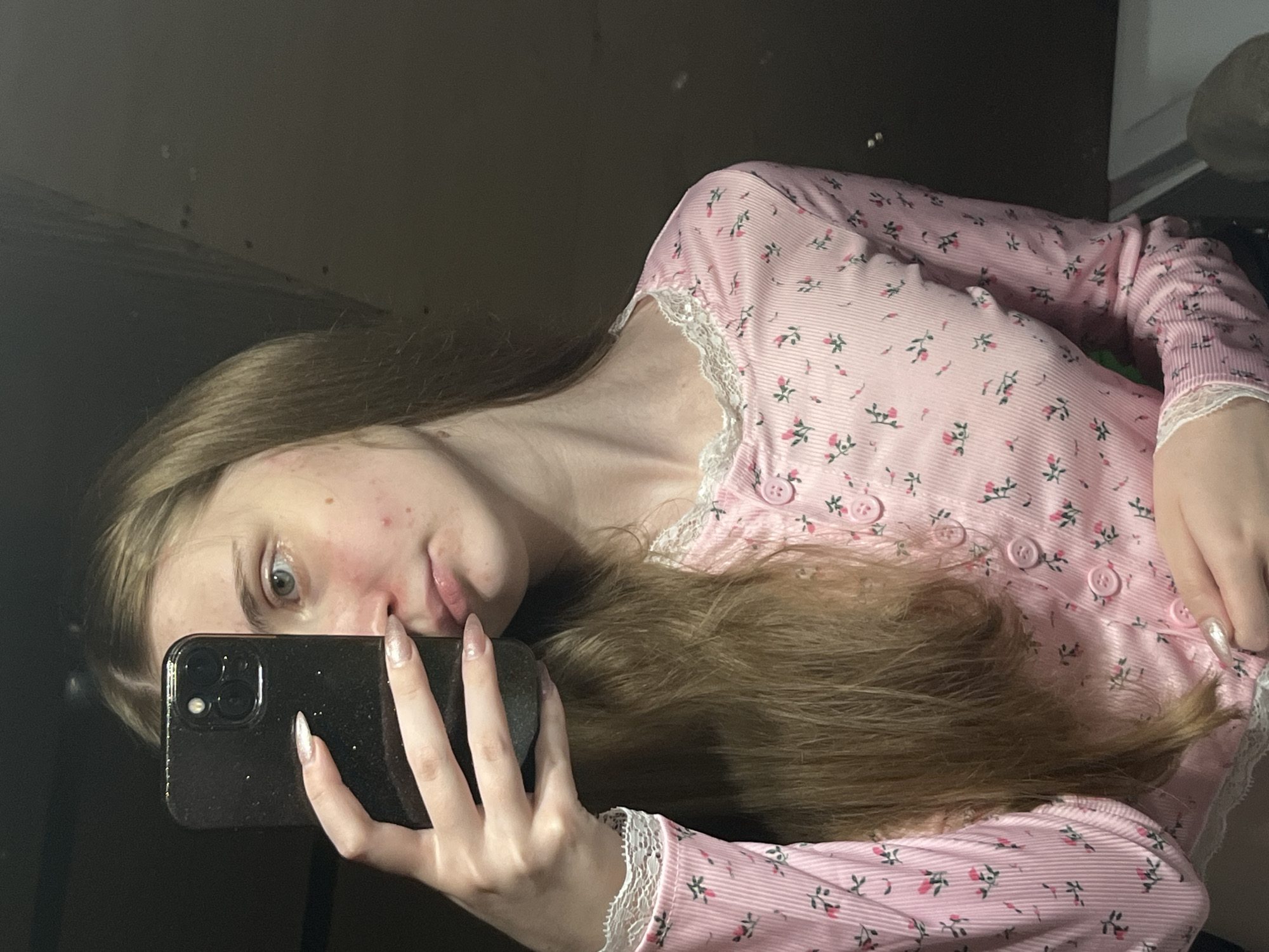 A young woman with light brown hair is taking a selfie in a dimly lit room. She is wearing a pink long-sleeved shirt with a floral pattern and white lace trim. Her left hand is holding up a dark-colored smartphone, partially obscuring her face. Her right hand is visible near her chest, with long, light-colored nails. The background appears to be a wall and possibly a ceiling, with some blurry details suggesting an indoor environment. There is no indication of the city of Ashkadar, Russia, or any specific location within it.