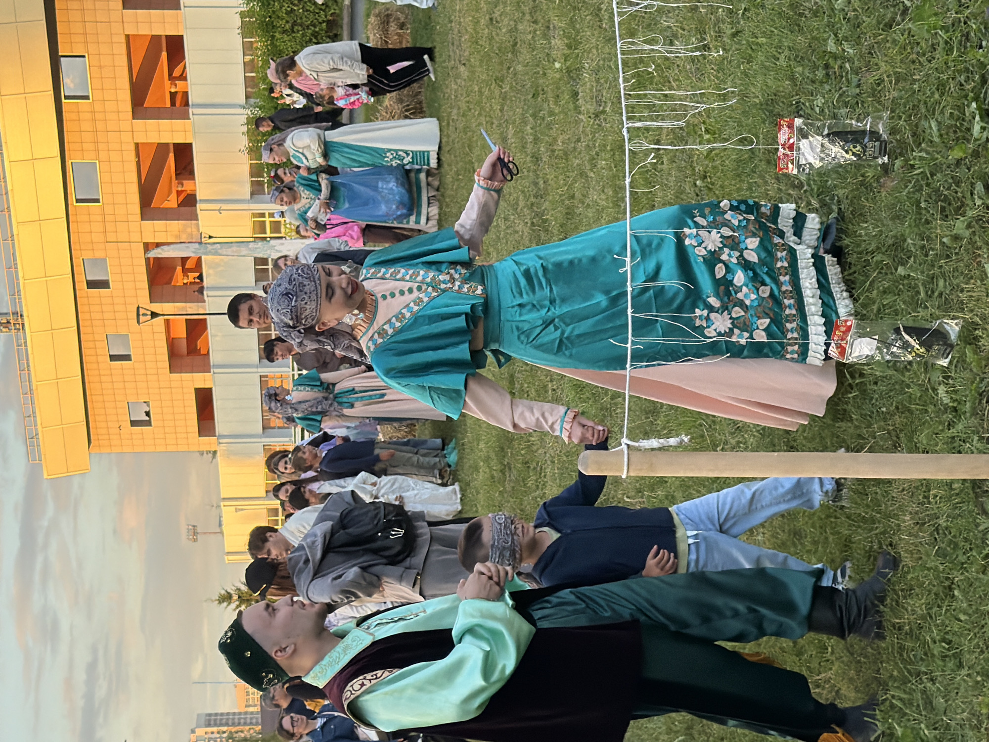 In the city of Unknown, Unknown, a group of people are gathered in an outdoor grassy area, possibly a park or festival grounds. In the foreground, a man and a woman, dressed in traditional ornate clothing, stand next to a young boy who is blindfolded and wearing a dark blue jacket and light blue jeans. The man is wearing a dark vest over a light green tunic and dark green pants, with a dark green cap. The woman is wearing a teal and pink traditional dress and a headscarf, and she is holding a pair of scissors. A white string with several white loops hangs horizontally across the scene, with two small red and white plastic-wrapped items hanging from it. In the background, a modern building with large windows and yellow trim is visible. Other people in the background are also dressed in various attire, some in traditional clothing and others in more casual modern clothes. The lighting suggests it is late afternoon or early evening.