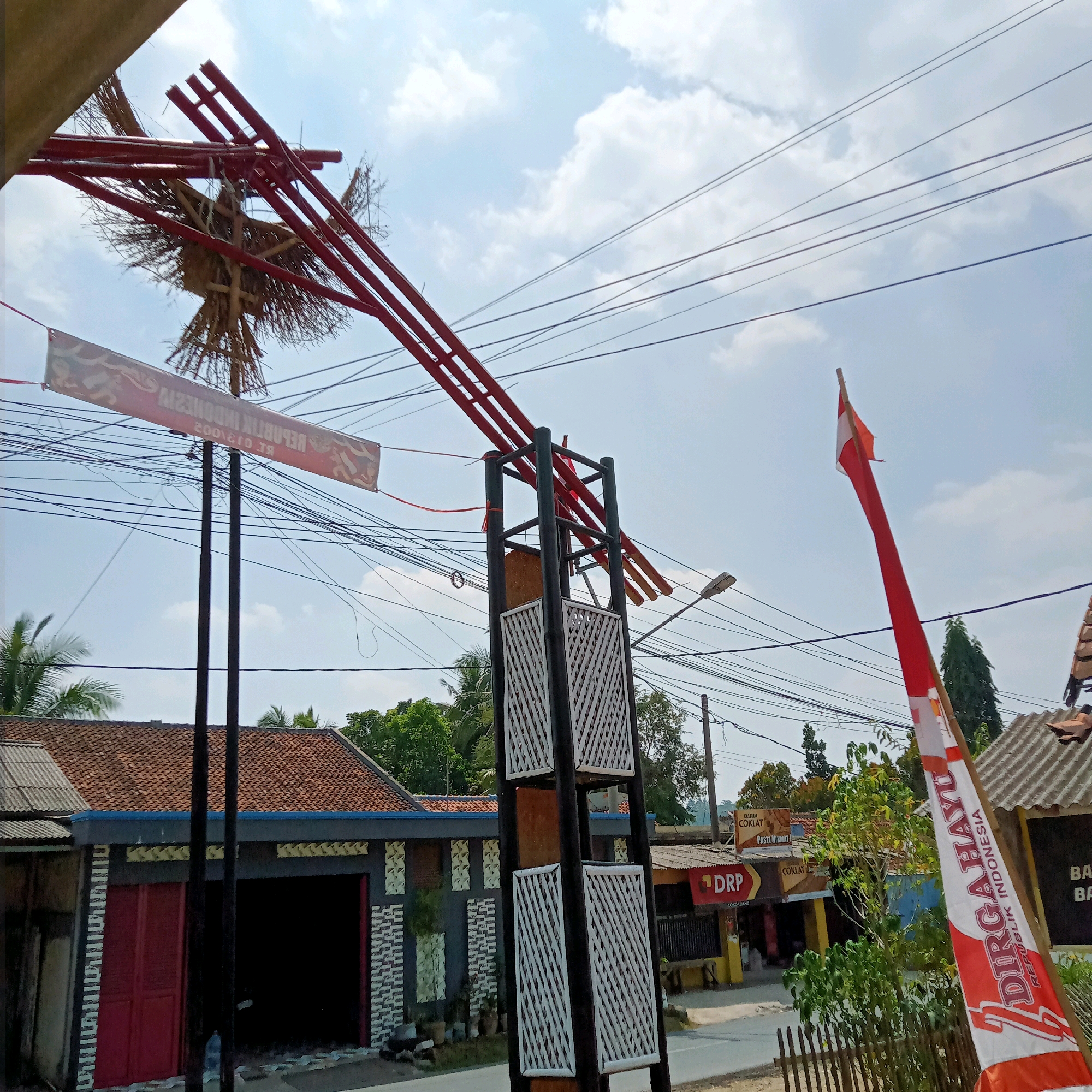 This is a medium-angle, eye-level exterior shot capturing a street scene in Purwakarta, Indonesia. Dominating the upper half of the frame is a bright blue sky with scattered white clouds. Several black electrical wires crisscross the sky, obscuring parts of the background.
In the foreground, a large, ornate red metal structure with a woven, bird-like decoration at its center extends from the top left of the frame. Below this structure, a horizontal red and white banner with text is hung. Two dark vertical posts support the banner. To the right, a tall Indonesian flag, red and white, waves in the breeze.
The middle ground shows a street lined with buildings. On the left, a building with a tiled roof and dark gray siding features a prominent red door and a large, open dark doorway. Decorative white and gray patterned panels adorn the facade. Further down the street on the right, a row of shops with various signs, including "DRP" and "COKLAT," are visible. A street lamp is also present on the right side of the road.
The background consists of lush green trees and more buildings with tiled roofs. A wooden fence runs along the bottom right of the image. The overall impression is a vibrant, somewhat cluttered, yet characteristic Indonesian streetscape under a clear sky.